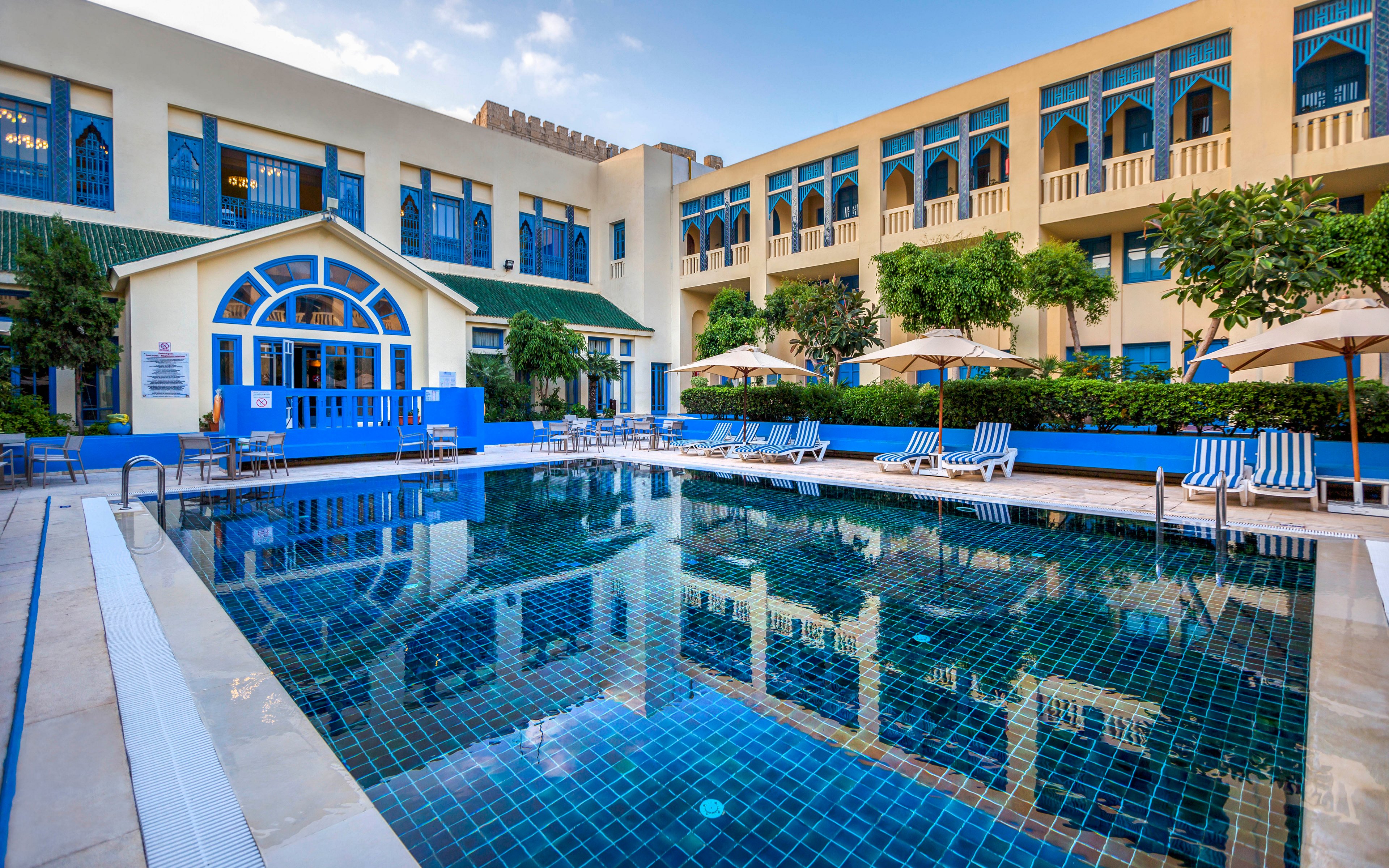 An outdoor swimming pool sits in the courtyard of a bright hotel or resort. The blue-tiled pool reflects the surrounding building with arched windows and balconies. Sun loungers and umbrellas line the poolside, suggesting a relaxing leisure area for guests. Green trees and clear skies add a fresh, inviting atmosphere to the scene.