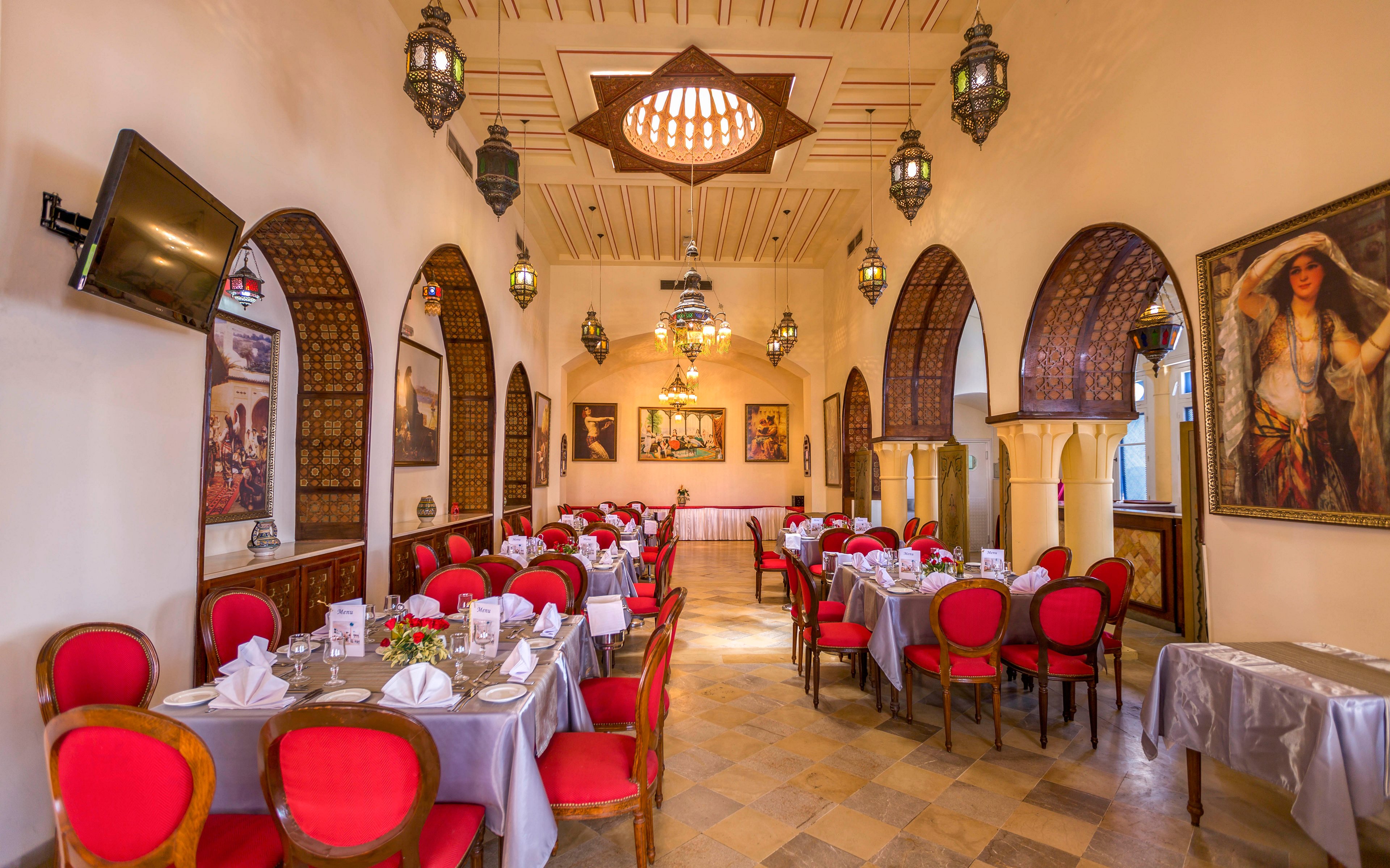 An ornate restaurant dining room is set with neatly arranged tables covered in white and gray linens. Red upholstered chairs surround the tables, which are decorated with folded napkins, glassware, and floral centerpieces. The space features arched doorways, carved wooden details, and hanging lantern-style lights that create a warm ambiance. Framed paintings and artwork adorn the walls, enhancing the traditional Middle Eastern aesthetic.