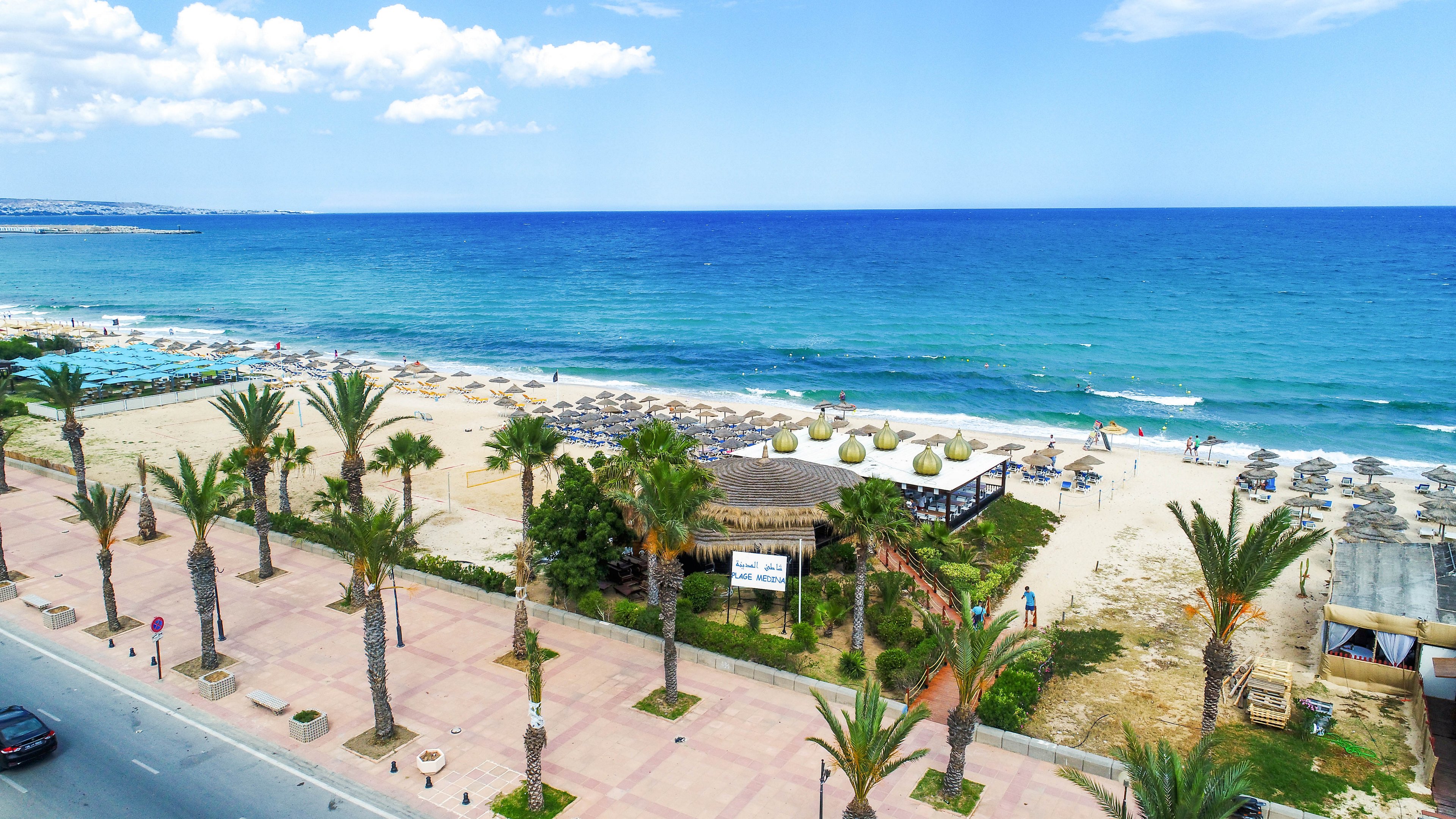 A wide sandy beach stretches along a bright blue sea under a partly cloudy sky. Rows of sun loungers and umbrellas line the shoreline, with a small beach bar or café nestled among palm trees. A paved promenade with palm trees runs parallel to the beach, separating it from the road. A few people walk along the sand and near the water, enjoying the coastal scenery.
