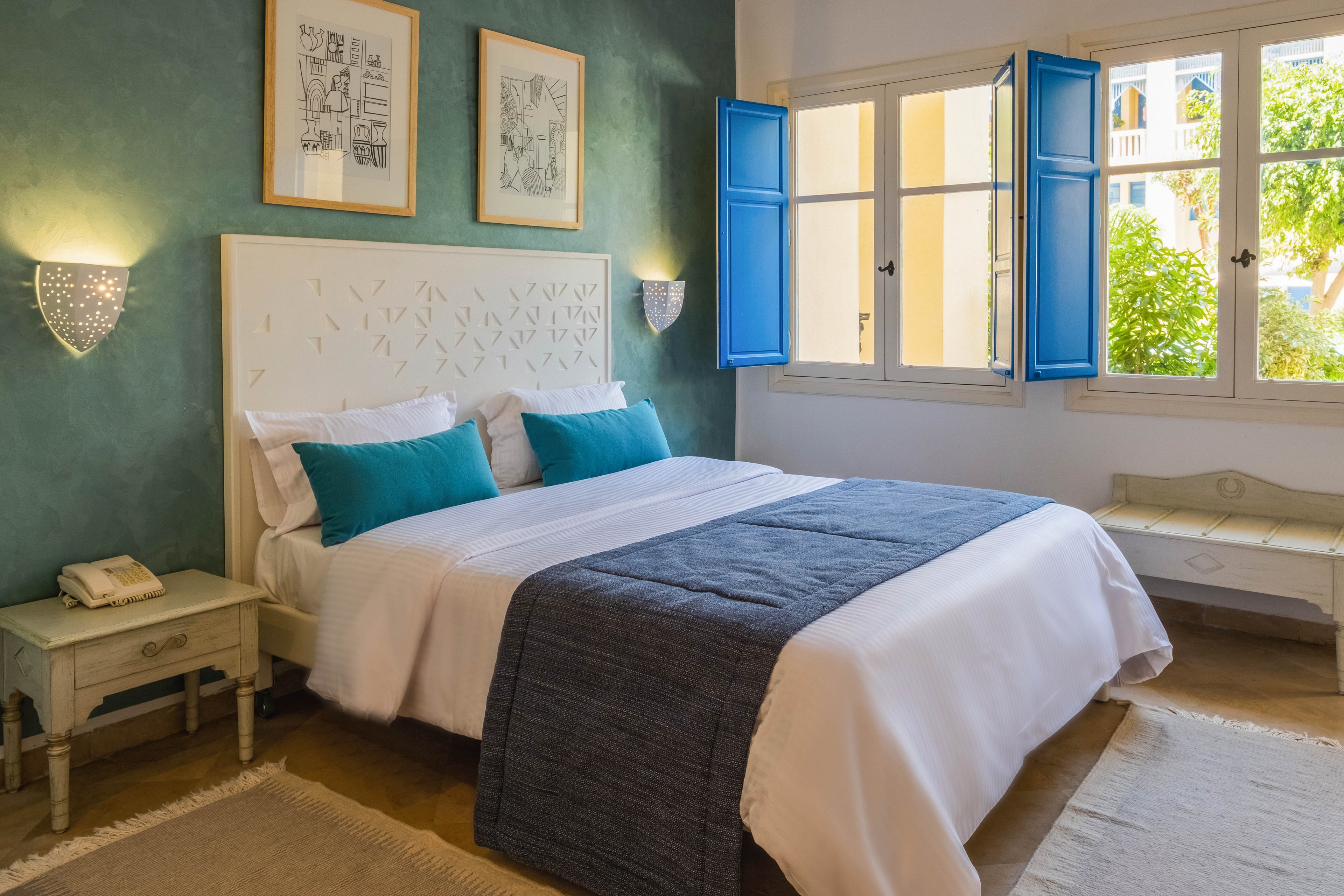 A neatly made double bed with white linens and teal cushions stands against a textured green wall in a bright bedroom. Blue shutters frame the open windows, letting natural light fill the room and revealing greenery outside. Matching bedside tables with lamps and a vintage telephone add a classic touch. A bench under the window and neutral rugs on the floor complete the calm, inviting atmosphere.