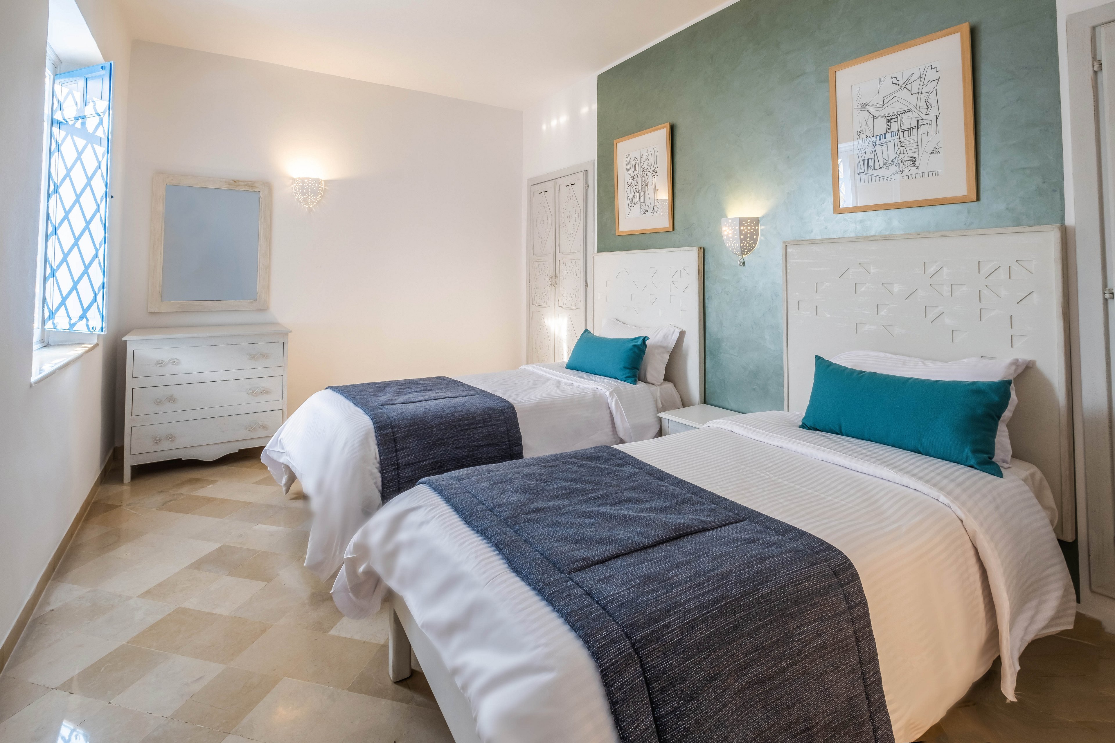 A bright bedroom features two neatly made twin beds with white linens, teal accent pillows, and dark blue throws. The room has a polished light-tiled floor and a soft green accent wall behind the headboards. Framed artwork hangs above the beds, and a white dresser with a mirror stands near a window with decorative bars. Wall sconces provide warm lighting, creating a clean and inviting atmosphere.