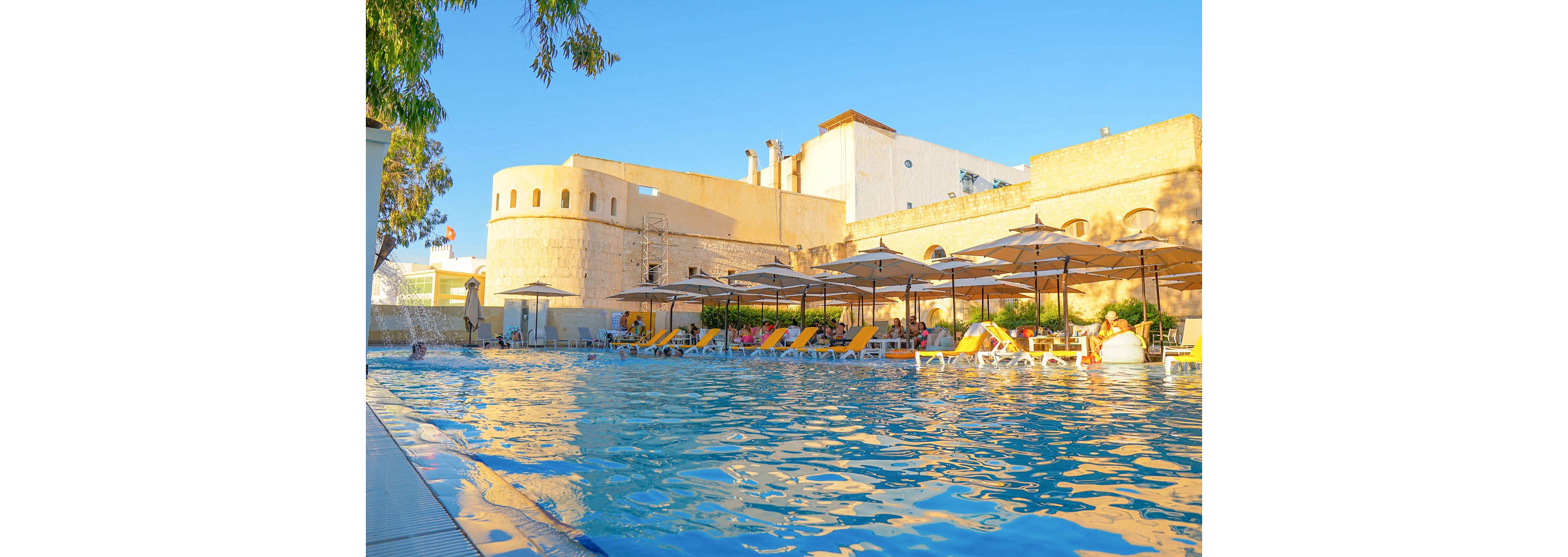 A bright outdoor swimming pool stretches across the foreground of a sunny resort scene. Guests relax on yellow lounge chairs under large umbrellas along the poolside. Behind them, tall beige stone walls and towers evoke a historic fortress or old city architecture. Clear blue water reflects the warm afternoon light, creating a tranquil vacation atmosphere. Trees frame the scene, adding greenery to the golden and blue color palette.