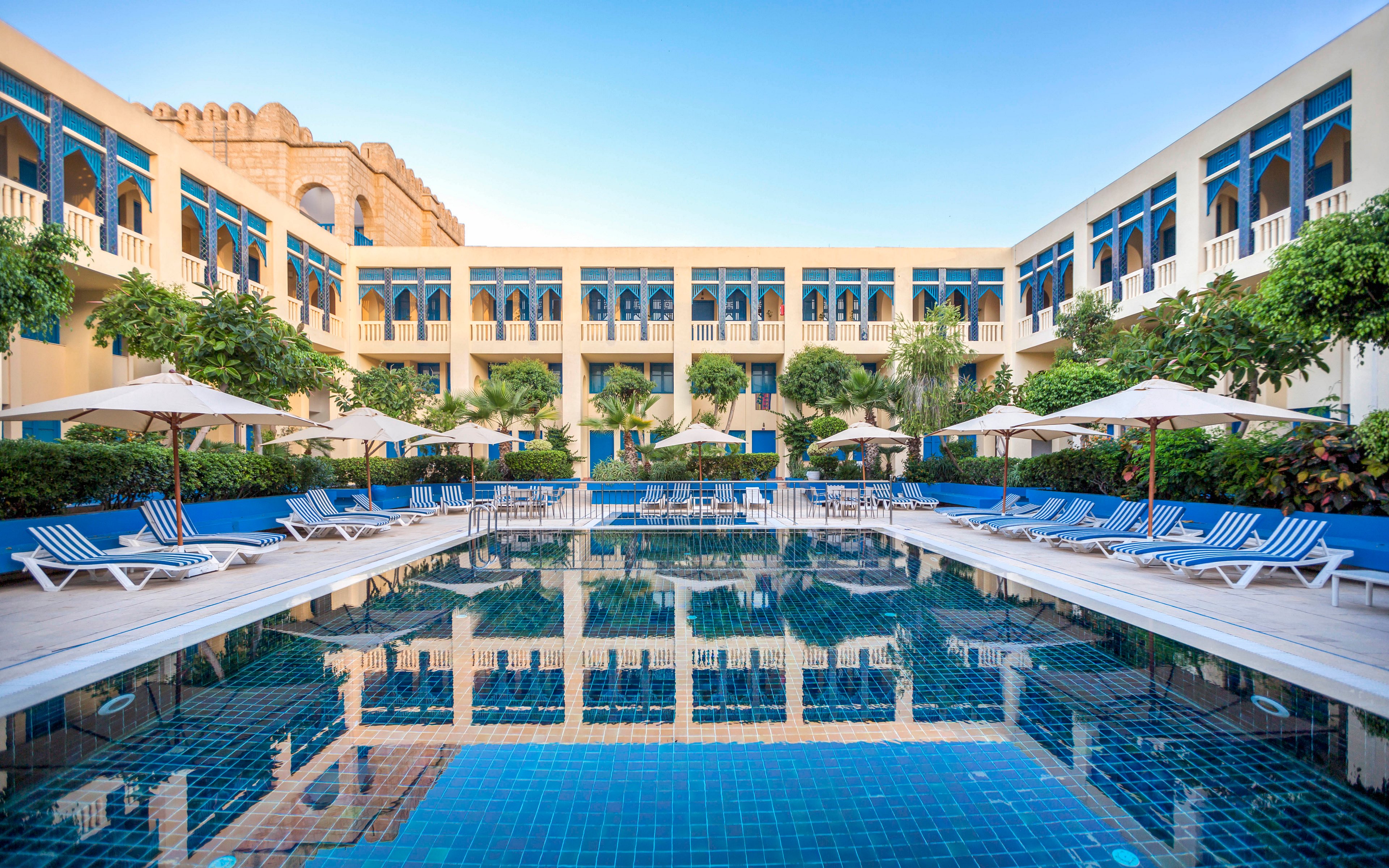 A large rectangular swimming pool sits at the center of a hotel courtyard. The calm water reflects the surrounding cream-colored building with blue window accents. Sun loungers and white umbrellas line both sides of the pool, creating a relaxing resort atmosphere. Green trees and shrubs add contrast to the blue tiles and clear sky above.