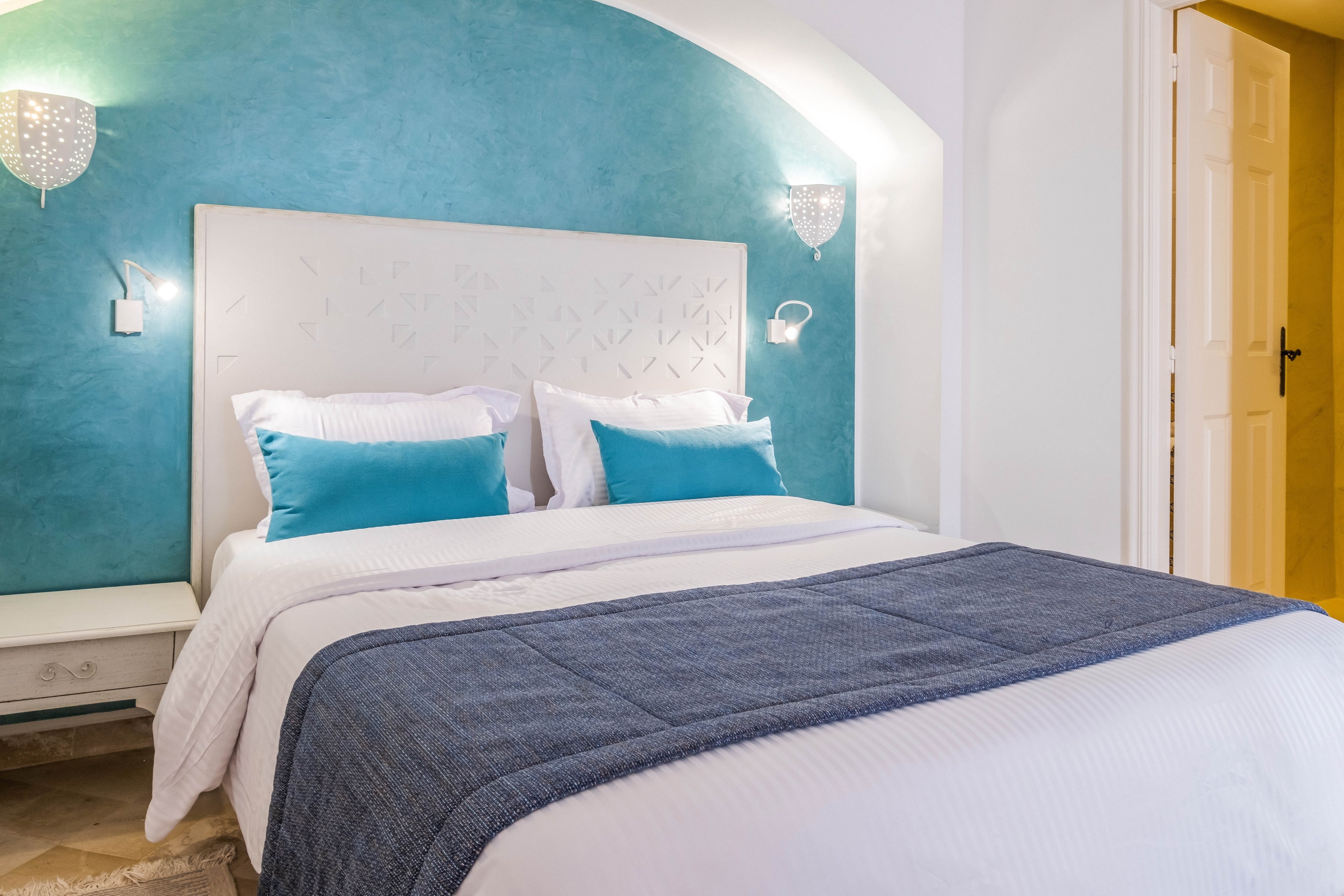 A neatly made double bed with white linens and teal accent pillows stands against a curved teal feature wall. Two wall-mounted lamps provide soft lighting on either side of the upholstered headboard. A bedside table with a drawer sits to the left, while an open doorway reveals a warmly lit adjacent room. The overall scene feels clean, inviting, and contemporary with a cool-toned color palette.