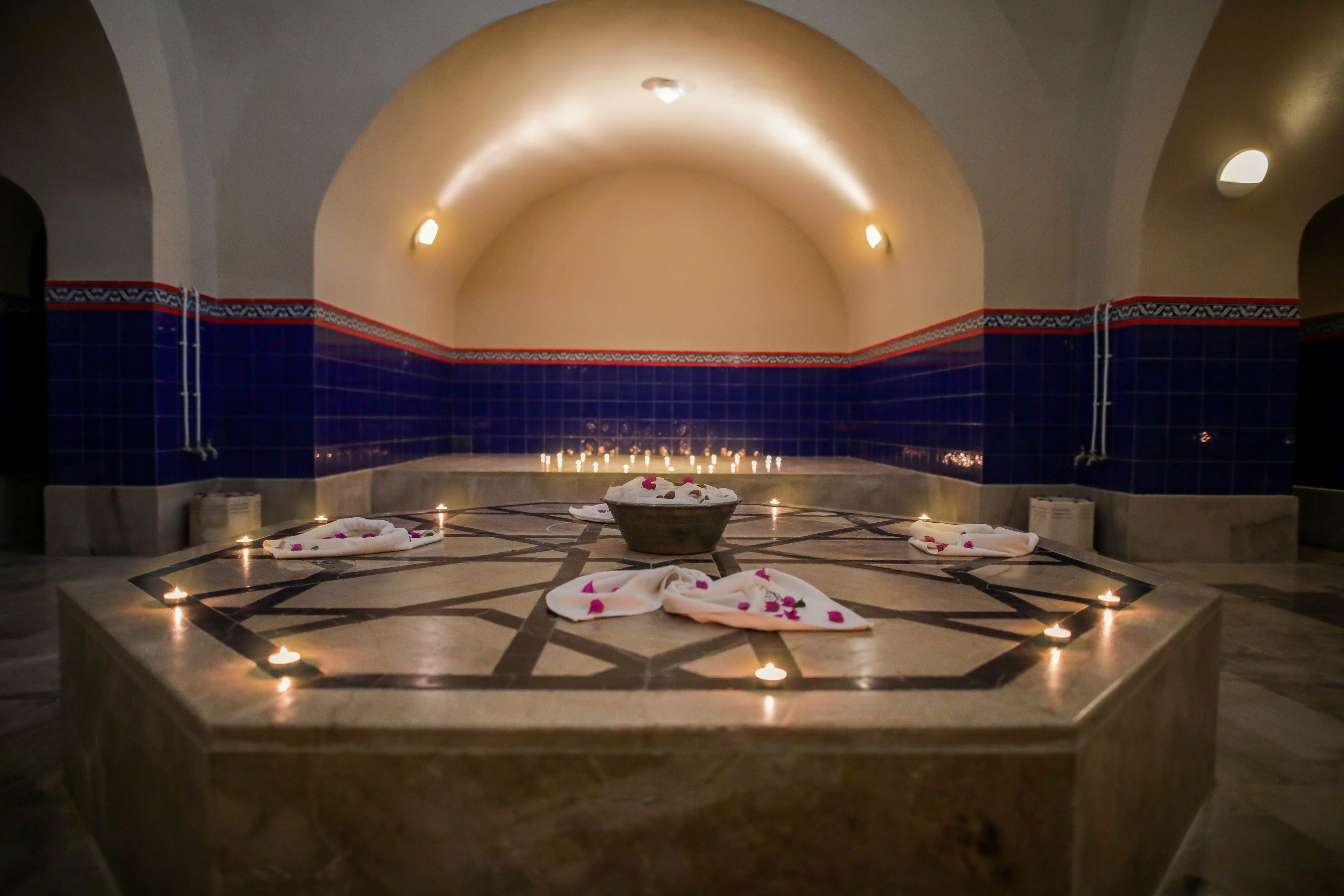 A serene spa hammam room features a central stone platform decorated with white towels and scattered flower petals. Small candles are arranged around the platform and along the walls, casting a warm, soft glow. The arched ceiling and deep blue tiled walls create a traditional bathhouse atmosphere. The overall scene suggests relaxation, wellness, and a tranquil retreat.