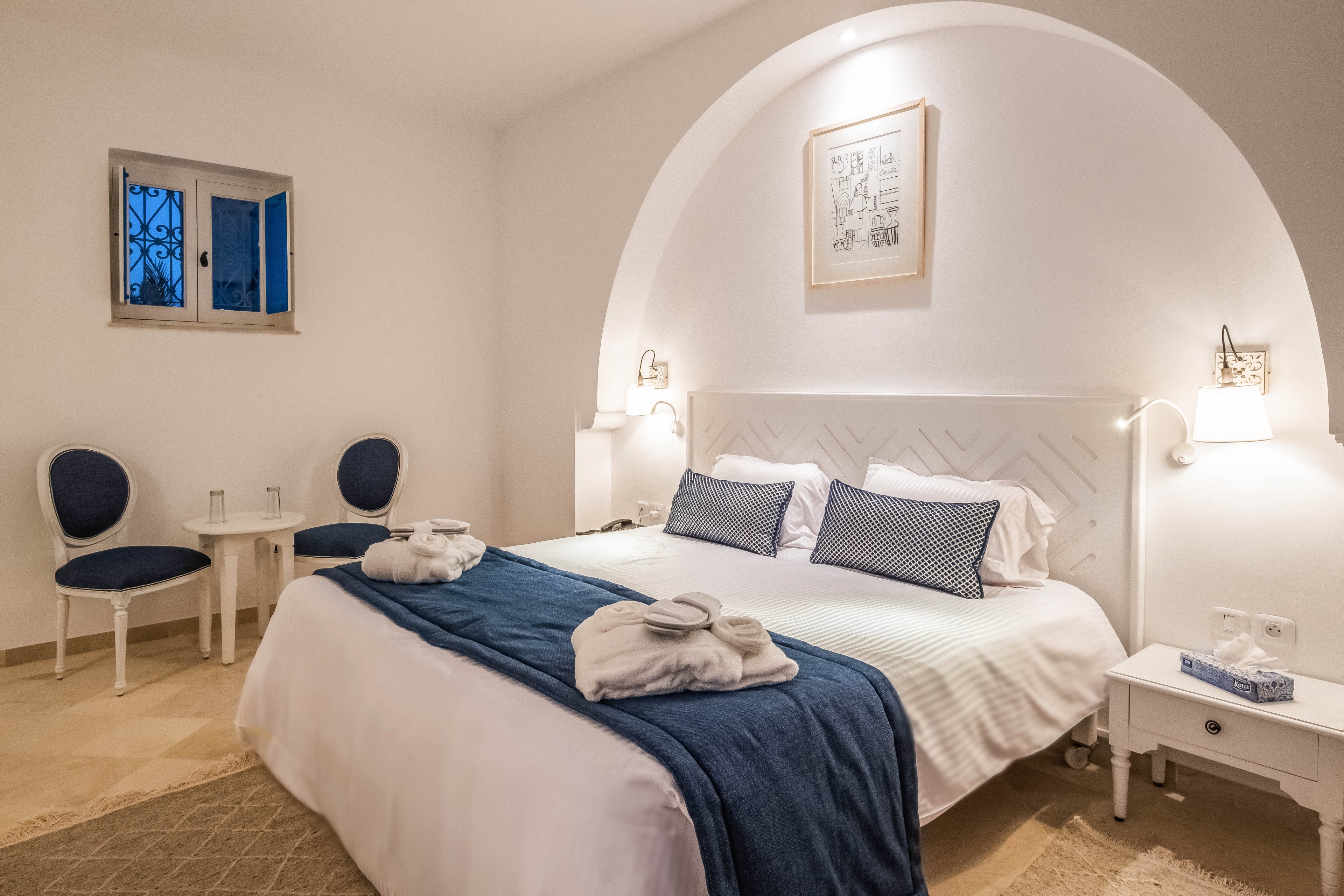 A neatly made double bed with white linens and navy blue accents is the focal point of this cozy bedroom. The room features soft, warm lighting, a small bedside table, and two upholstered chairs near a window with decorative bars. Rolled white towels are placed on the bed, suggesting a hotel or guest room setting. The overall design is minimalist and clean, with neutral walls and subtle artwork above the headboard.