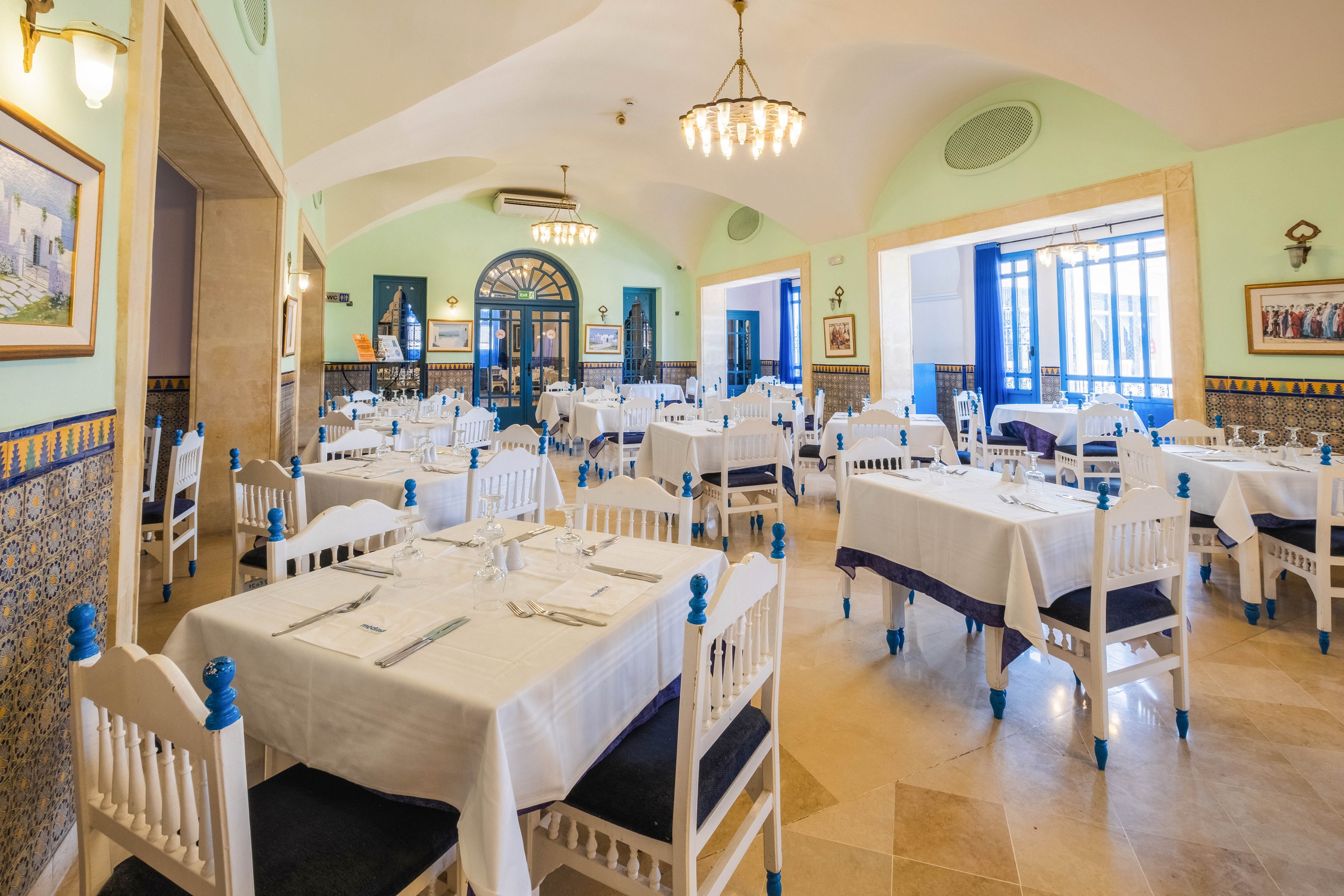 A spacious restaurant dining room is set with neatly arranged white tables and chairs. The room features arched ceilings, tiled walls, and large windows letting in natural light. Blue accents on the chairs and decor create a Mediterranean coastal feel. The tables are prepared with cutlery and glasses, but no diners are present, giving a calm and orderly atmosphere.