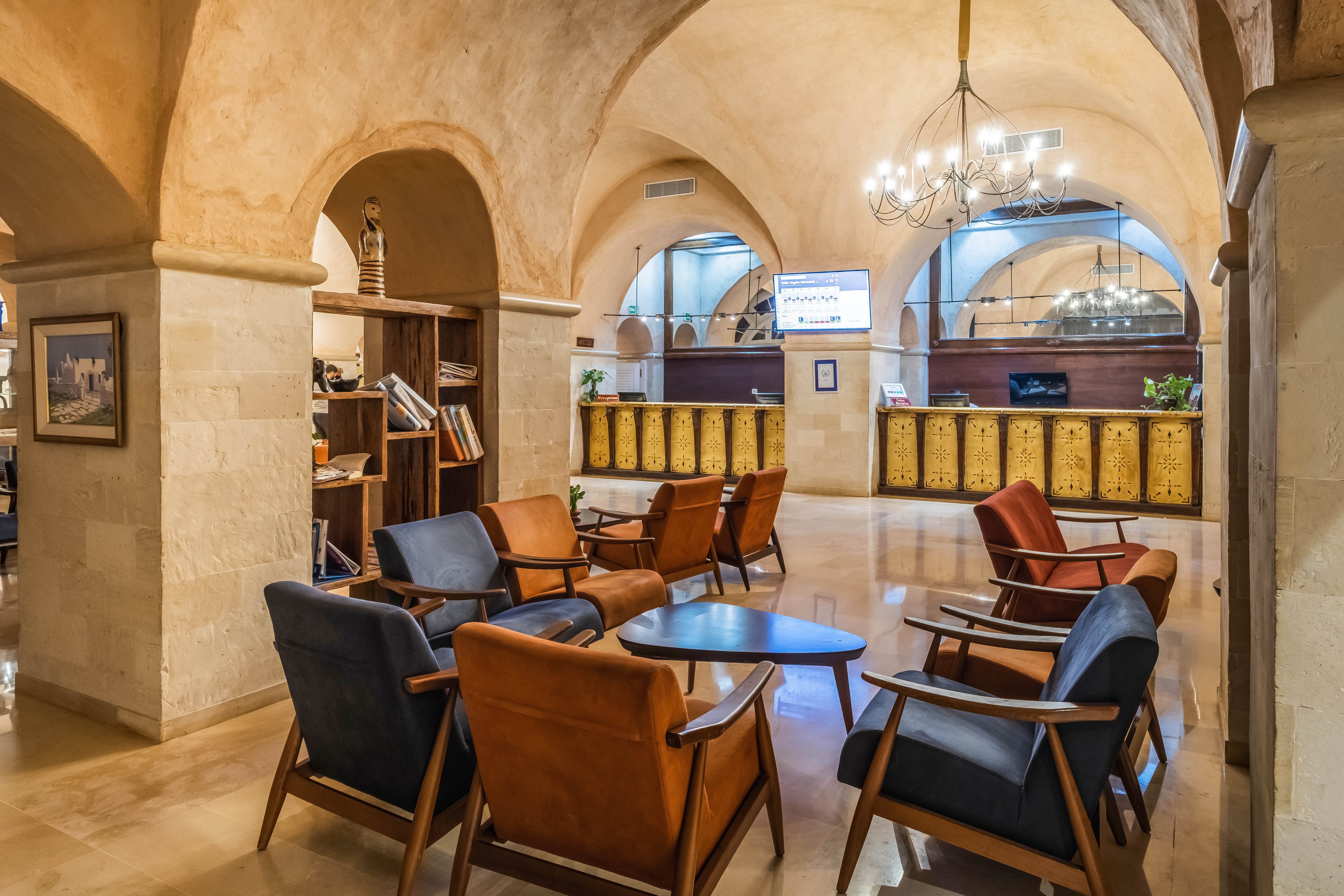 A stylish hotel lobby features mid-century modern armchairs arranged around small tables beneath high vaulted stone ceilings. The reception desk stretches across the back of the room, with decorative panels and warm lighting creating an inviting atmosphere. A bookshelf with various items and books stands to the side, adding a cozy lounge feel. A chandelier and recessed lighting highlight the polished floor and architectural arches, giving the space a refined yet comfortable ambiance.