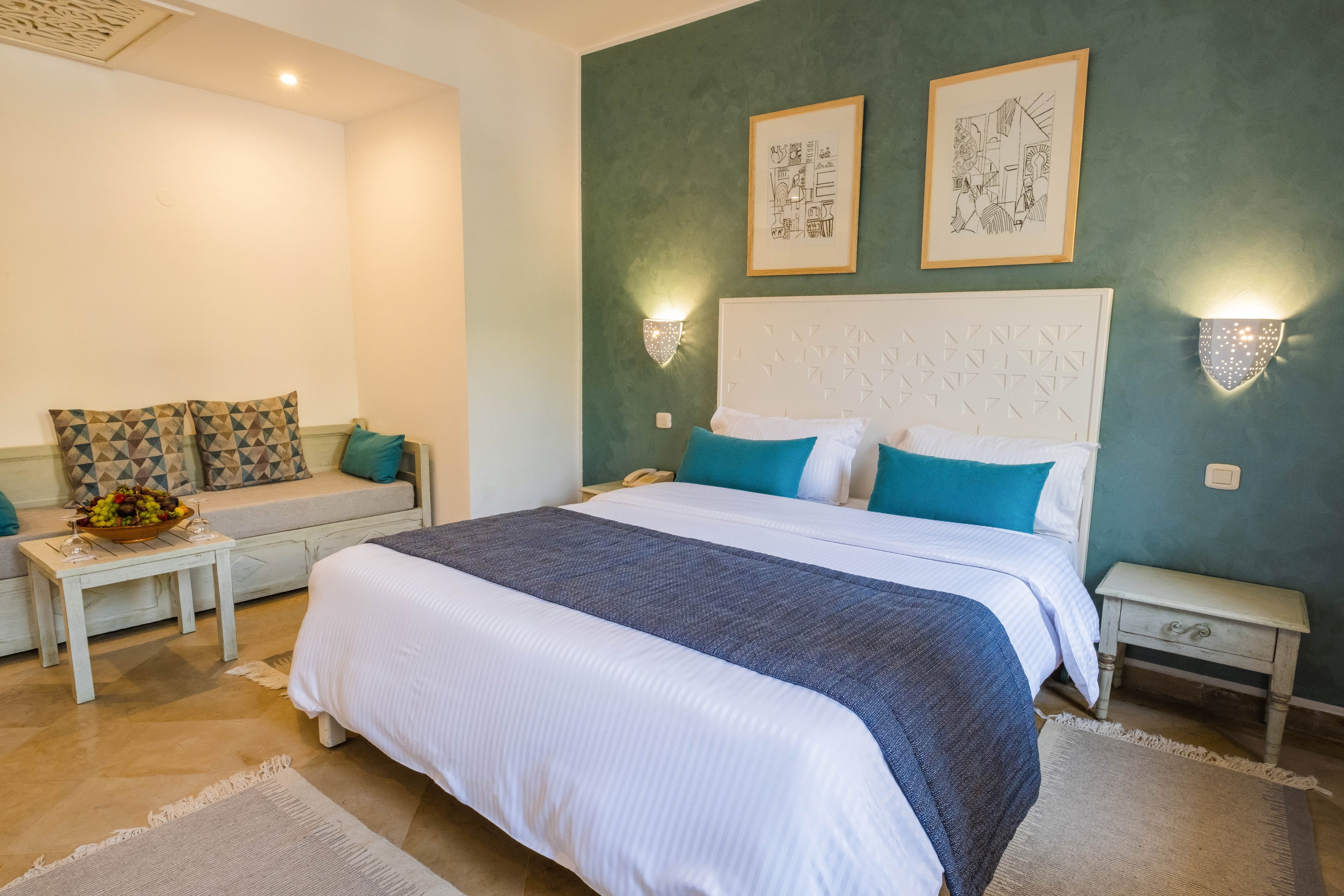 A neatly made double bed with white linens and teal accent pillows is the focal point of this modern bedroom. The room features a green accent wall with two framed artworks above the headboard and wall-mounted sconces providing warm light. To the side, a cushioned sofa nook with patterned pillows and a small table holding a fruit arrangement adds a comfortable seating area. Neutral rugs and light-colored furniture create a calm, inviting atmosphere suitable for a hotel or guest room.