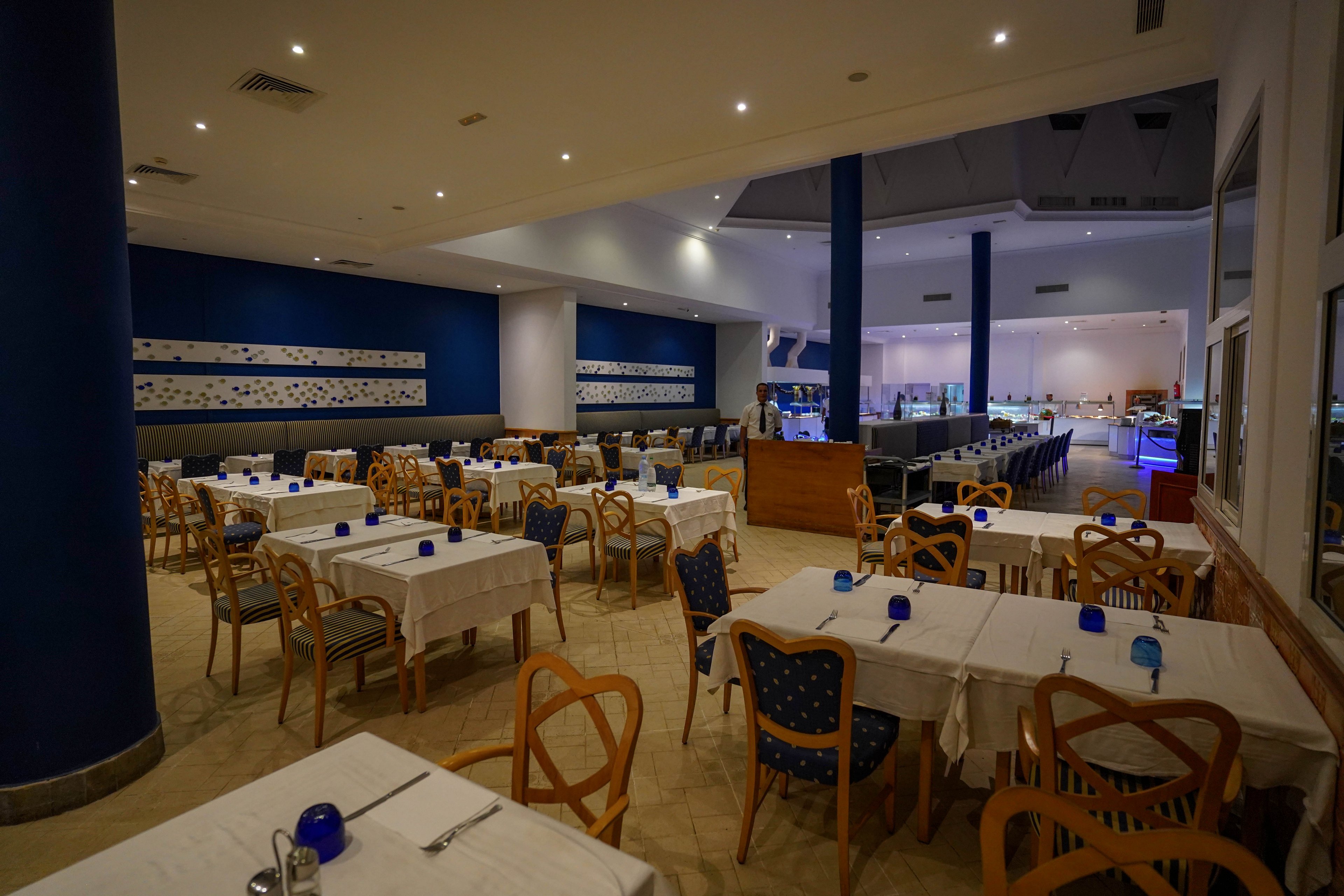A spacious restaurant dining room is set with neatly arranged tables and wooden chairs. White tablecloths and simple place settings with cutlery and blue glass candle holders create a clean, modern look. The space features blue accent walls, columns, and upholstered chairs, with buffet counters visible in the background. Warm indoor lighting and the absence of people give the scene a calm, quiet atmosphere.