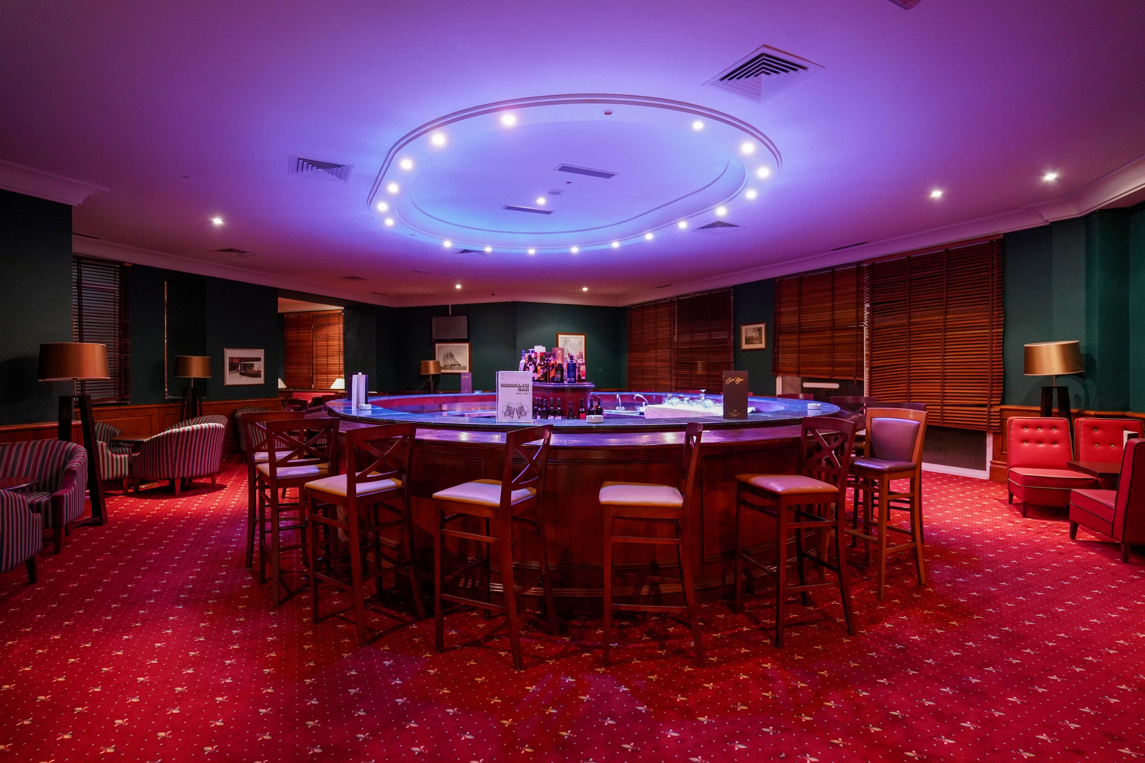 A circular bar counter stands in the center of an empty lounge, surrounded by wooden bar stools. The room features deep red carpet with a patterned design and dark green walls. Purple ceiling lights cast a soft glow over the bar area, highlighting bottles and glassware. Comfortable armchairs and small tables line the perimeter, creating an inviting yet quiet atmosphere.