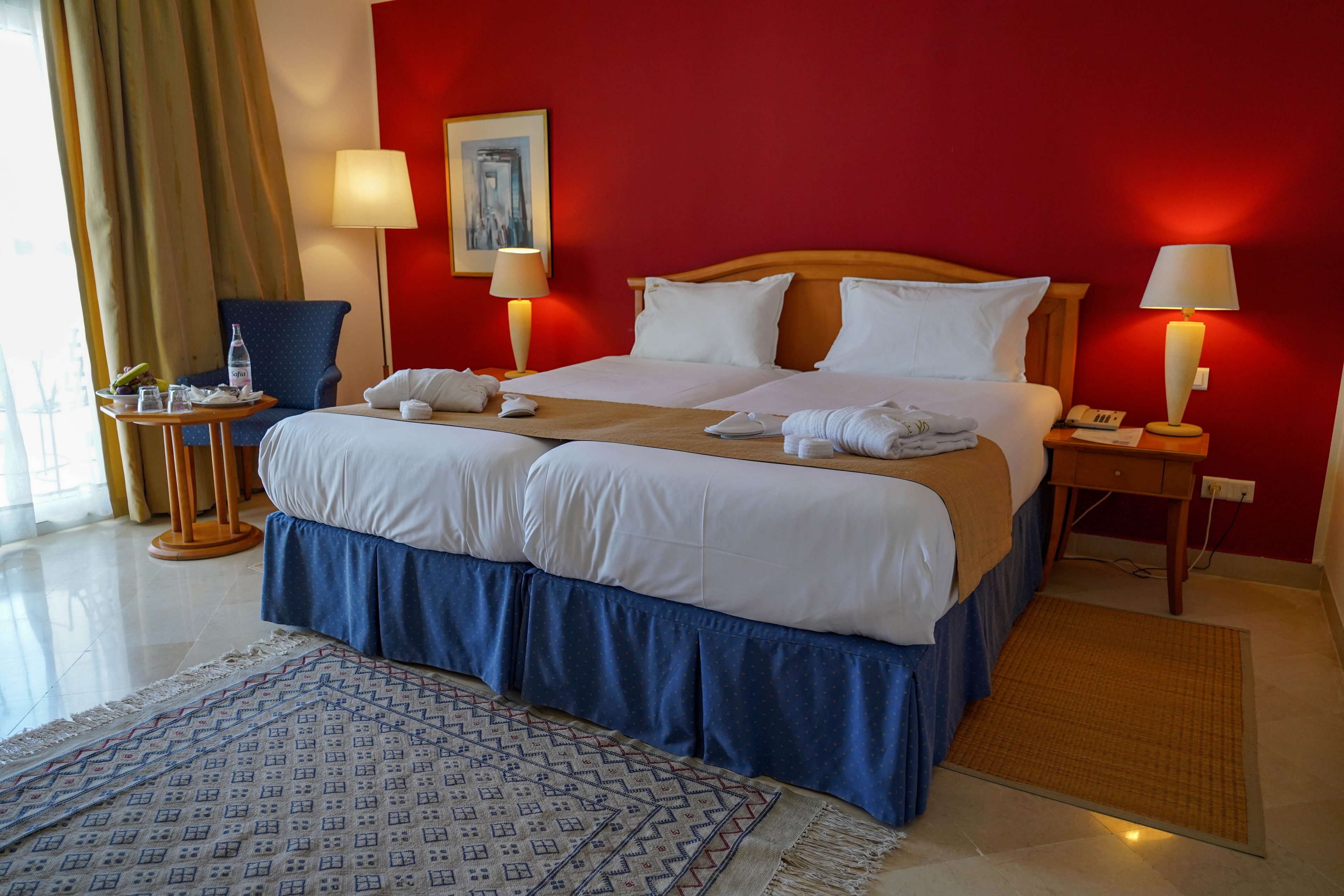 A neatly made twin hotel bed with white linens and tan runners stands against a bold red accent wall. Two bedside tables with matching lamps frame the bed, creating a warm and inviting atmosphere. A small seating area with a round table, chair, and bottled water is visible near the window with long curtains. The polished floor, woven rug, and framed artwork add to the comfortable, contemporary hotel room setting.