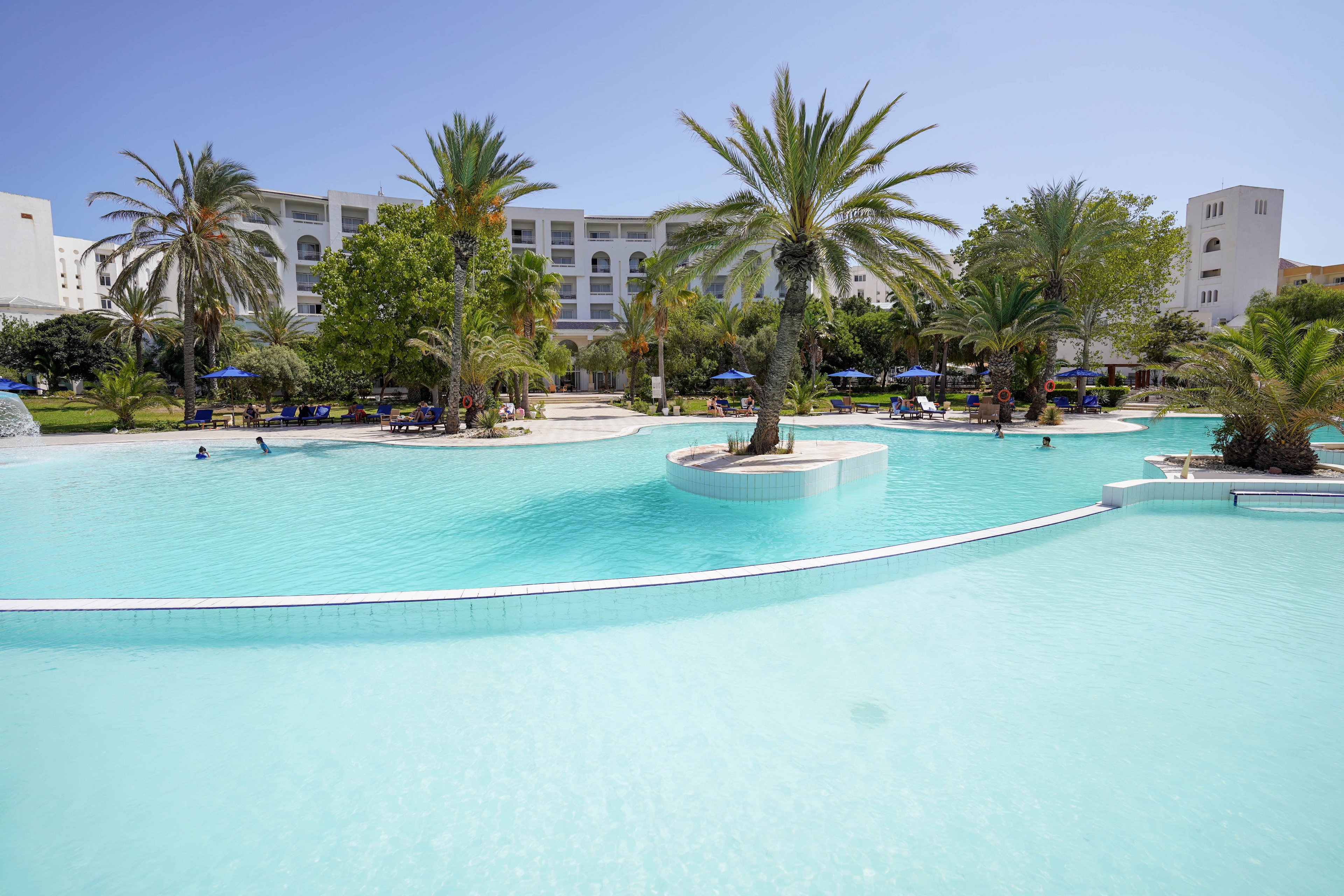 A large, bright blue swimming pool stretches across the foreground of a resort complex. Tall palm trees and lush greenery surround the water, creating a tropical atmosphere. A white multi-story hotel building stands in the background under a clear blue sky. Guests relax on sun loungers and under umbrellas along the poolside, suggesting a warm vacation setting.
