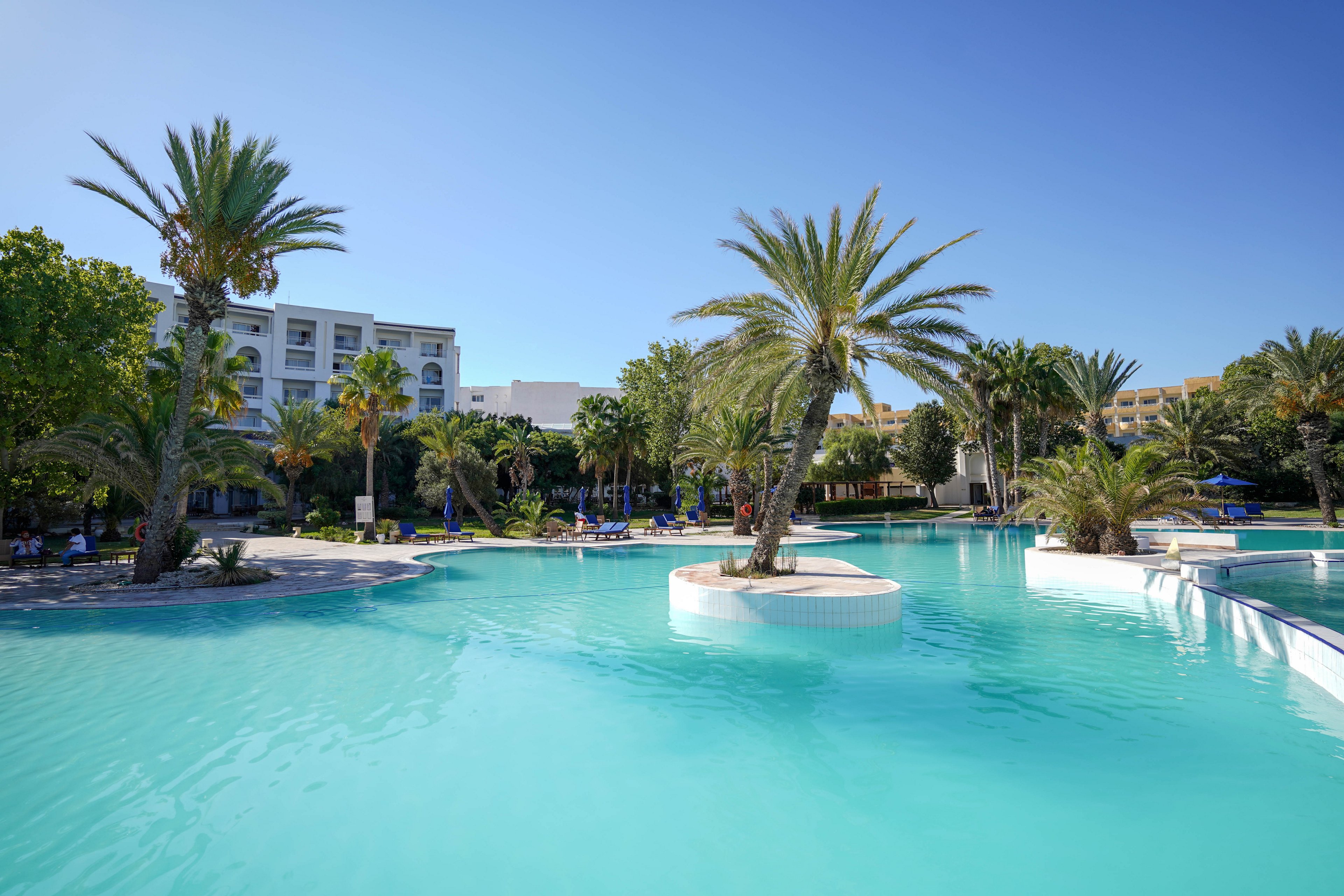 A large turquoise swimming pool stretches across the foreground of a tropical resort. Tall palm trees rise from small islands within the pool and along its edges. A white multi-story hotel building and other resort structures are visible in the background under a clear blue sky. The scene suggests a sunny, relaxing vacation atmosphere with calm water and lush greenery.