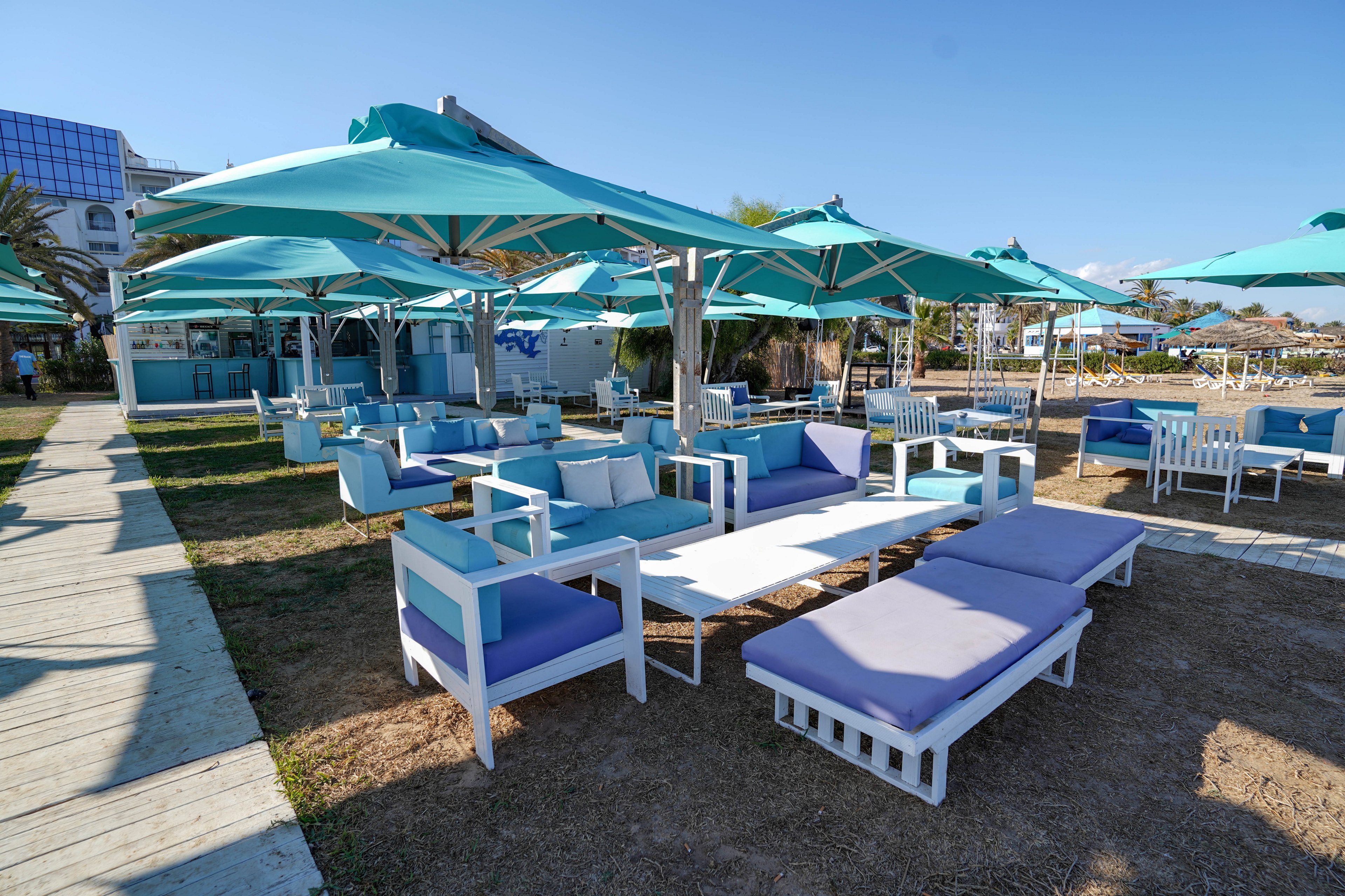 An open-air beach lounge area features modern white and turquoise seating under large teal umbrellas. Cushioned sofas, armchairs, and low tables are arranged on a mix of wooden decking and dry grass. The scene is bright and sunny with clear blue skies and a relaxed coastal atmosphere. In the background, more seating and structures suggest a seaside resort or beach club.