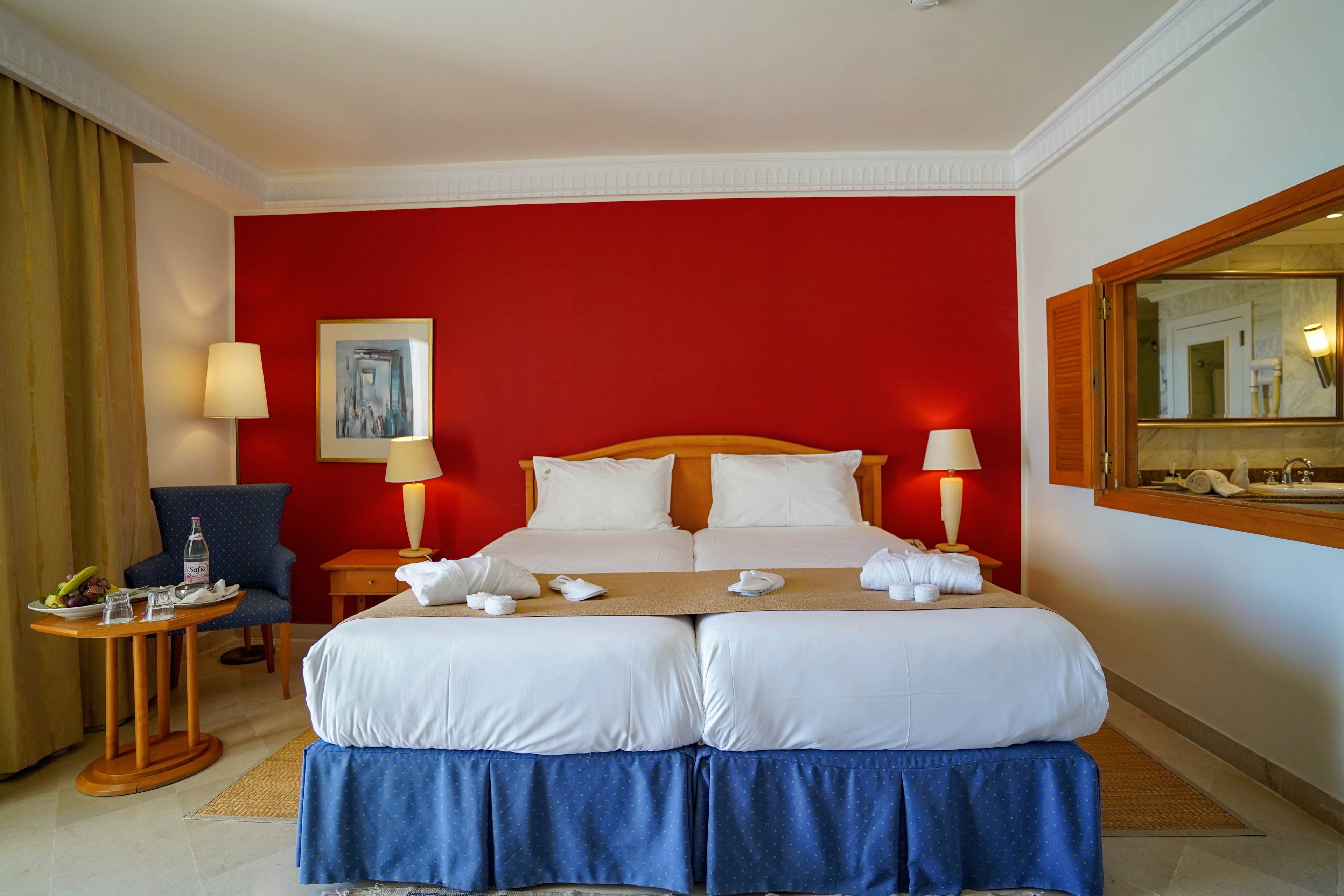 A neatly made double bed with white linens and blue bed skirt stands against a bold red accent wall in a modern hotel room. Two bedside tables with matching lamps frame the bed, and folded towels are arranged on the blanket. A small round table with a bottle of water, glass, and fruit sits near a blue armchair by the window with beige curtains. A framed artwork and a large wall mirror add decorative touches to the clean, contemporary space.