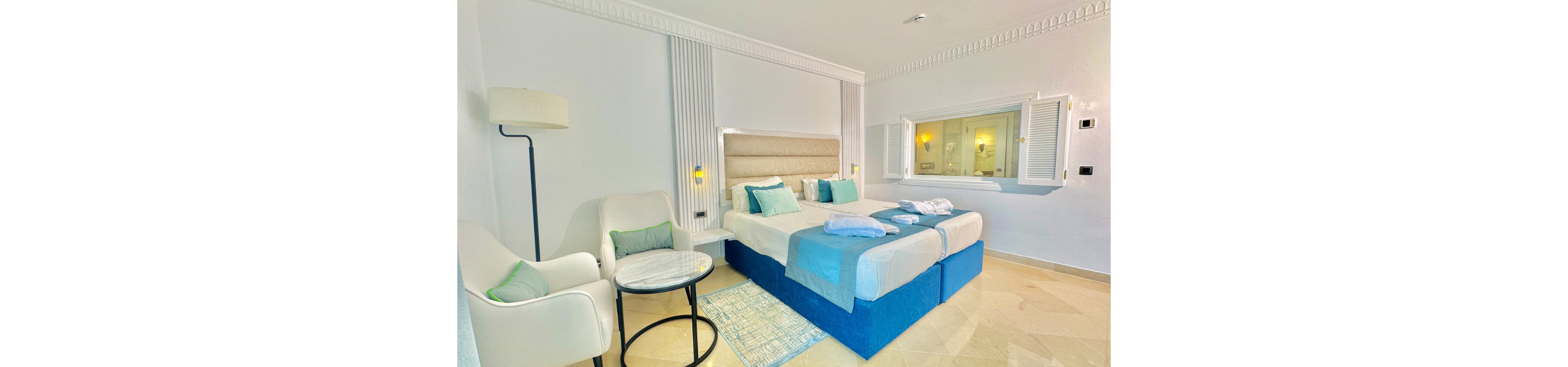 A bright, modern hotel bedroom features two twin beds pushed together with blue bases and coordinated bedding. Soft teal and white pillows and throws add color to the neutral walls and floor. A cozy seating area with an armchair, floor lamp, and small round table sits beside the bed. A large interior window opens to the bathroom, enhancing the airy, spacious feel of the room.