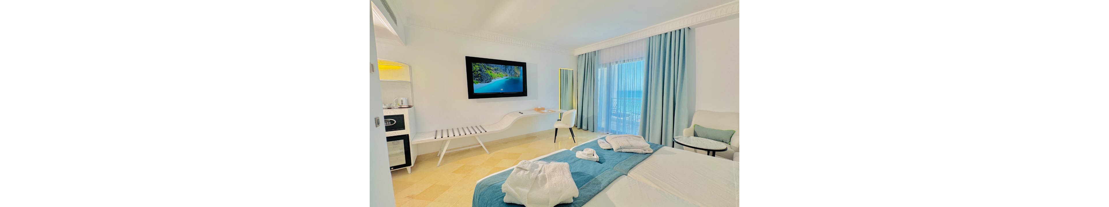 A modern hotel room features a neatly made bed with folded white bathrobes on a teal runner. A large flat-screen television on the wall displays a tropical beach scene, adding to the vacation atmosphere. Light blue curtains frame a glass door that opens to a balcony with an ocean view. Minimalist white furniture, including a lounge bench, desk, and armchair, completes the clean and airy interior.