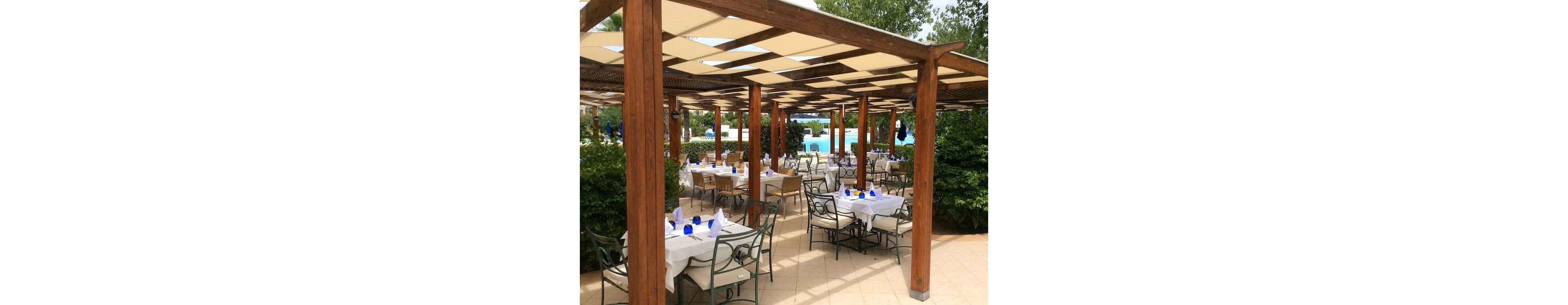 An elegant outdoor restaurant is set beneath a wooden pergola with a checkered canopy providing shade. Tables are neatly arranged with white tablecloths, napkins, and blue glassware, creating a refined dining atmosphere. The terrace overlooks a bright blue swimming pool surrounded by greenery and trees. The scene suggests a relaxed, resort-style setting prepared for guests.