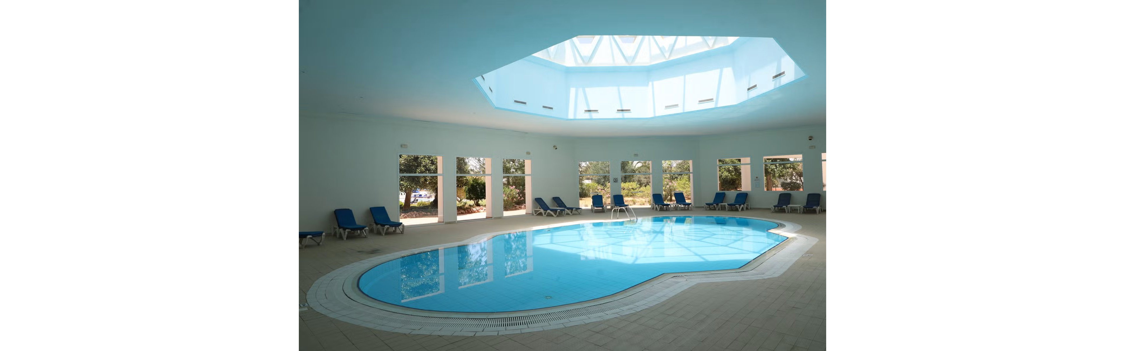 An indoor swimming pool sits at the center of a spacious, light-filled room. A large geometric skylight in the ceiling lets natural daylight flood the area. Blue lounge chairs line the tiled perimeter of the pool, facing large windows that reveal greenery outside. The scene feels calm and inviting, suggesting a relaxing leisure or spa environment.