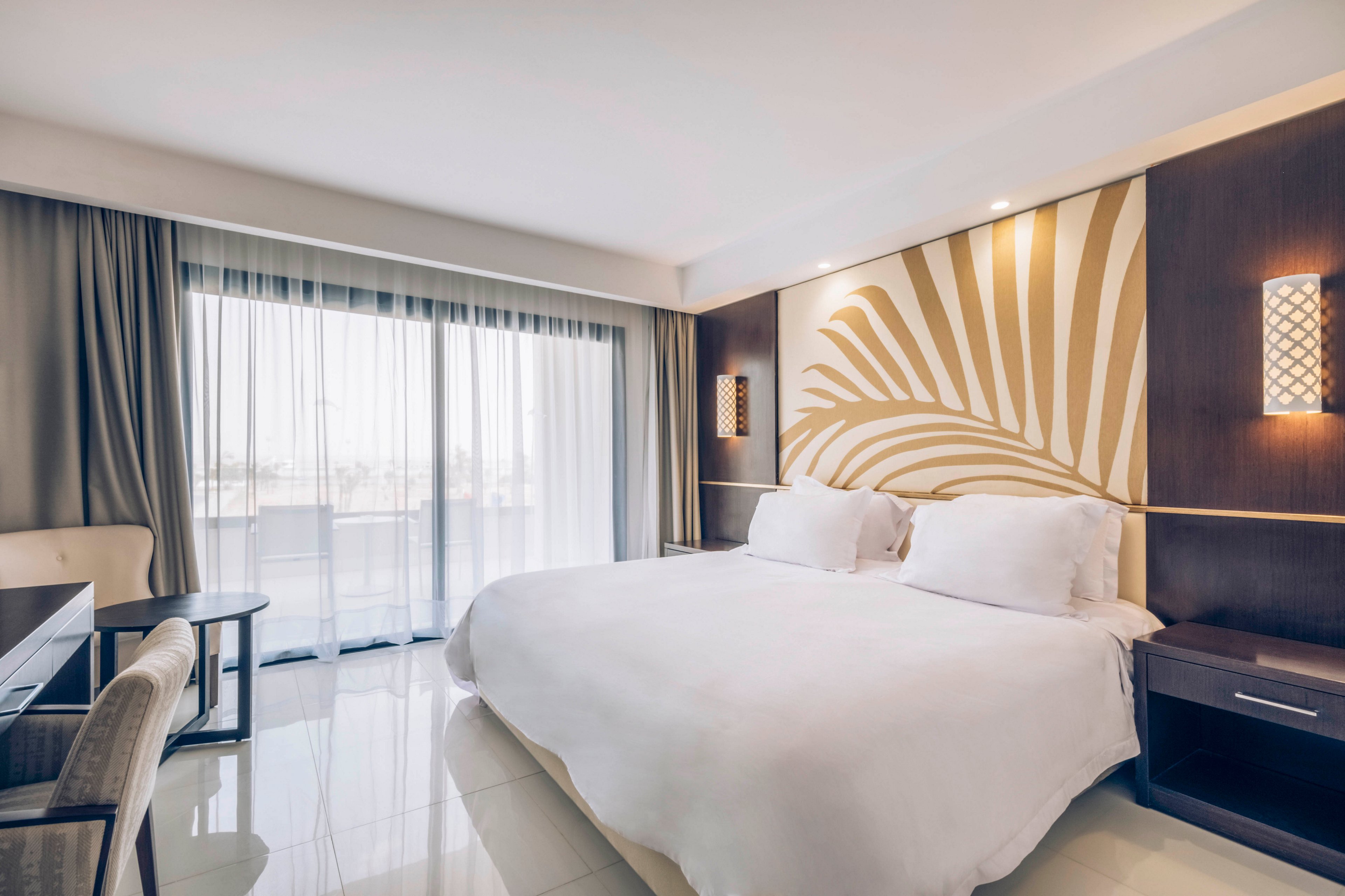A spacious modern hotel room features a large white bed with plush pillows against a decorative headboard. Warm wall sconces illuminate the room, highlighting the contemporary wood paneling and abstract leaf design. Floor-to-ceiling windows with sheer curtains open to a balcony, filling the space with natural light. A desk and chair sit to the side, creating a comfortable area for work or relaxation.