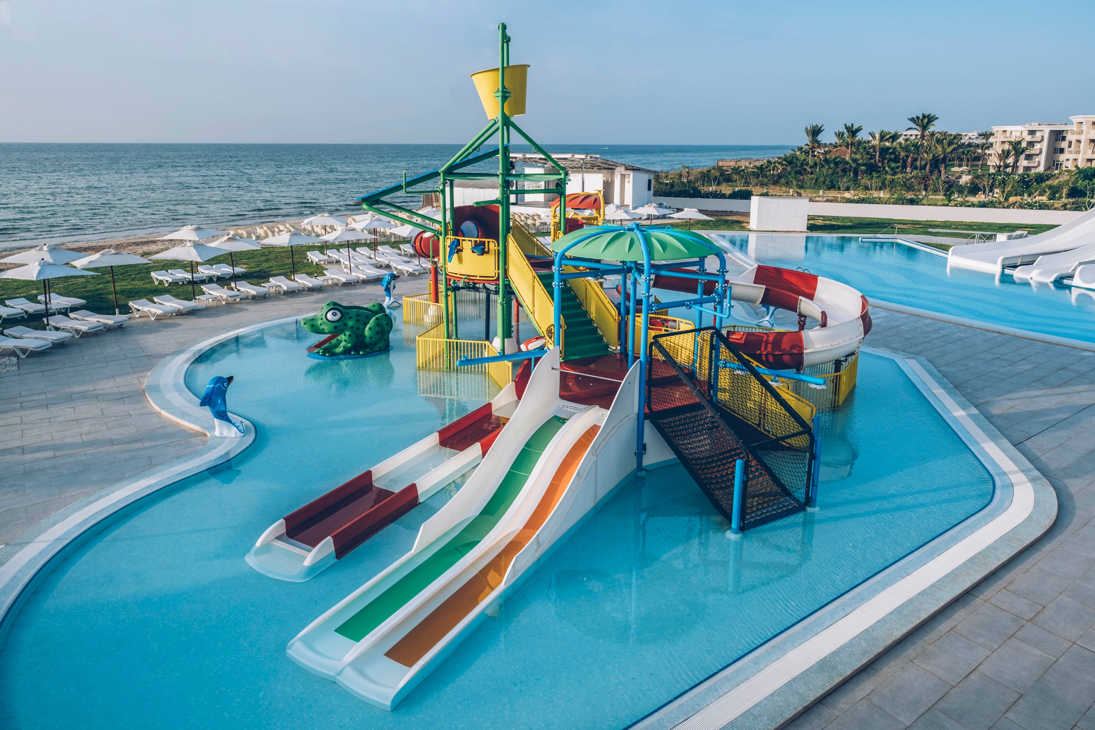 A colorful children's water playground with multiple slides sits in a shallow pool at a seaside resort. The structure features bright red, yellow, green, blue, and white elements, including tunnels, stairs, and splash areas. Lounge chairs and umbrellas line the pool deck, with the ocean and palm trees visible in the background. The scene suggests a family-friendly vacation destination focused on fun and relaxation by the water.
