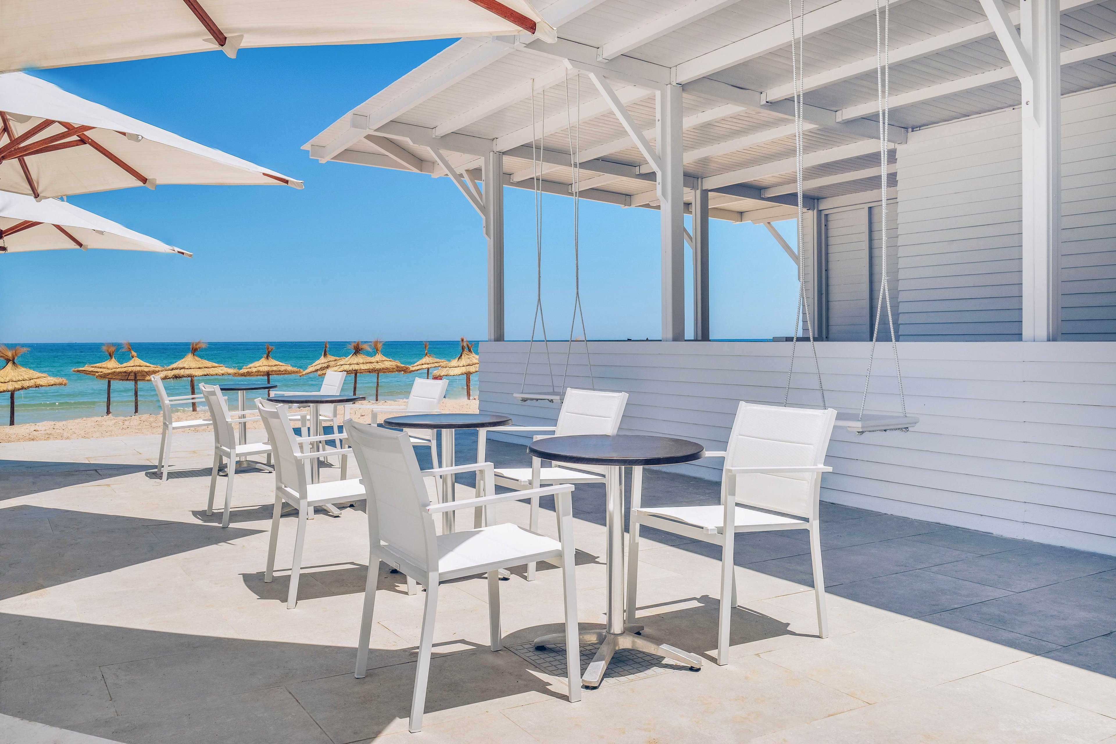 An open-air beachfront cafe terrace features white tables and chairs under a minimalist pergola. Wooden swings hang from the ceiling beside the seating area, adding a playful touch. In the background, straw parasols line the sandy beach with turquoise sea and clear blue sky. The bright sunlight and clean white surfaces create a fresh, summery atmosphere.