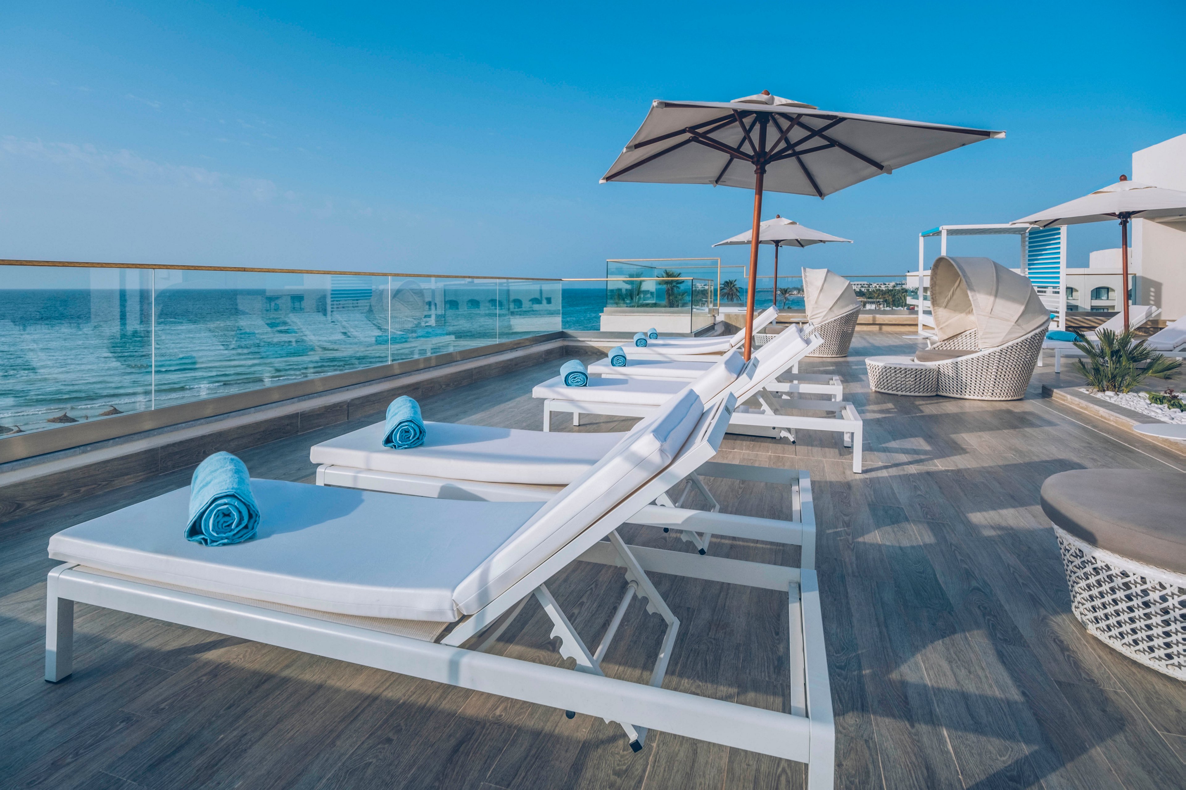 A row of white sun loungers with rolled blue towels lines a modern rooftop deck overlooking the ocean. Large umbrellas provide shade while glass railings offer an unobstructed view of the sea and sky. The wooden deck and contemporary outdoor furniture create a clean, upscale resort atmosphere. Calm blue tones and bright daylight emphasize a relaxing vacation setting.
