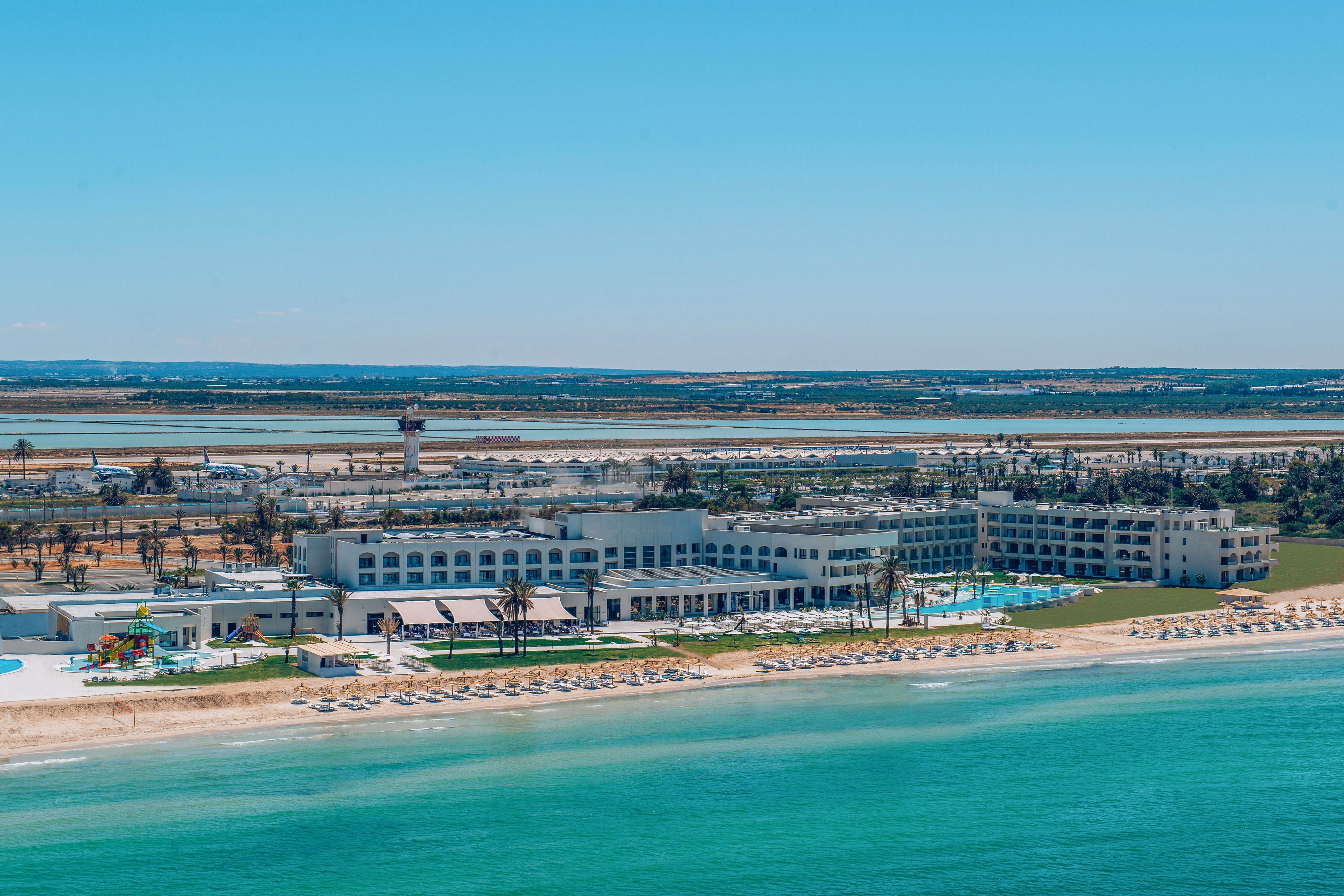 A large beachfront resort stretches along a sandy shoreline beside clear turquoise water. Multiple white buildings with balconies surround several outdoor swimming pools and palm-lined walkways. Rows of sun loungers and umbrellas are neatly arranged on the beach facing the sea. In the background, flat coastal land and a distant control tower are visible under a bright blue sky.