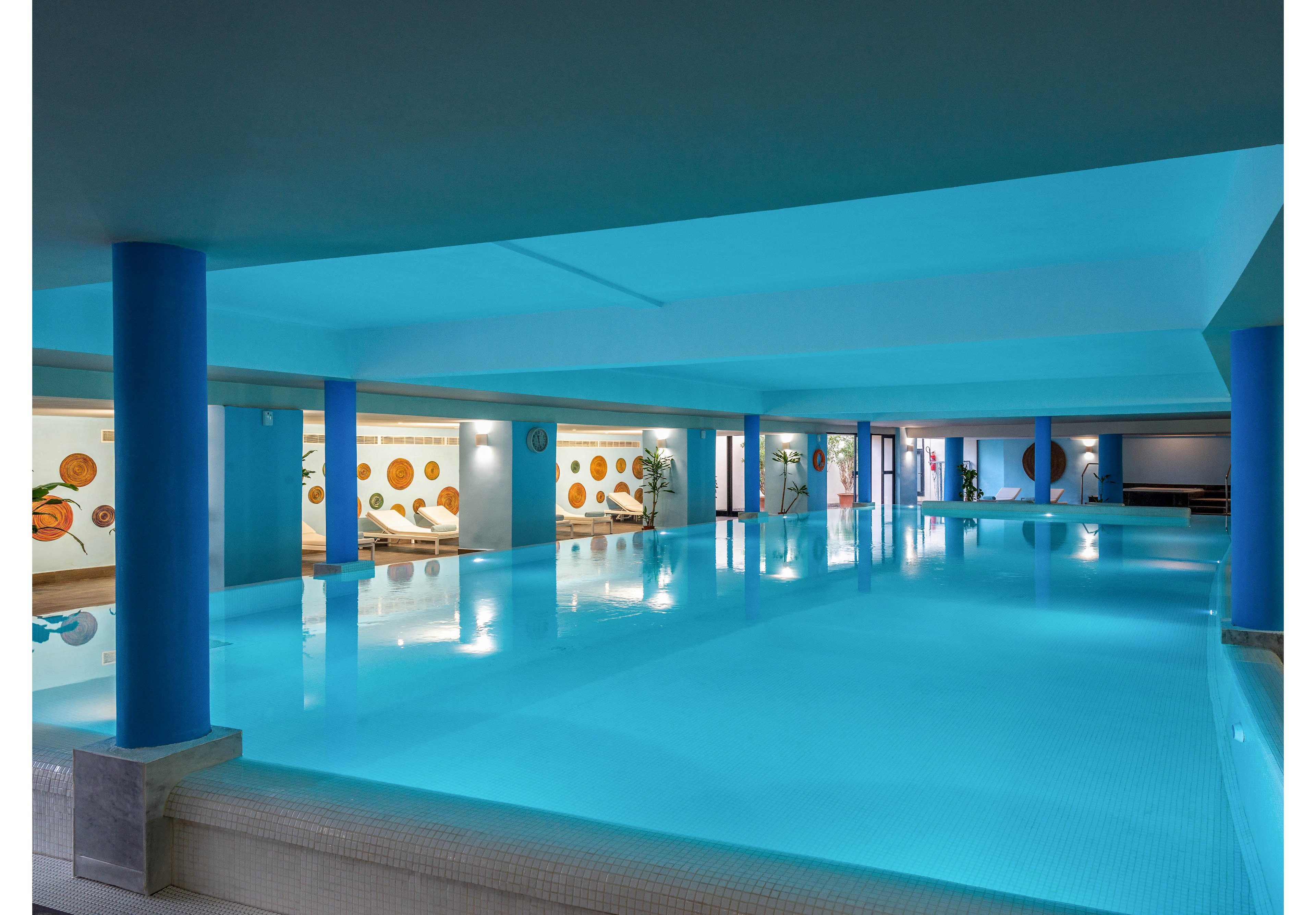 Spacious indoor swimming pool with calm, clear blue water dominates the scene. The pool is surrounded by blue structural columns and a low ceiling, creating a sleek, modern atmosphere. Warm wall art and recessed lighting add contrast along the perimeter. No people are present, emphasizing a quiet, relaxing environment suitable for leisure or exercise.