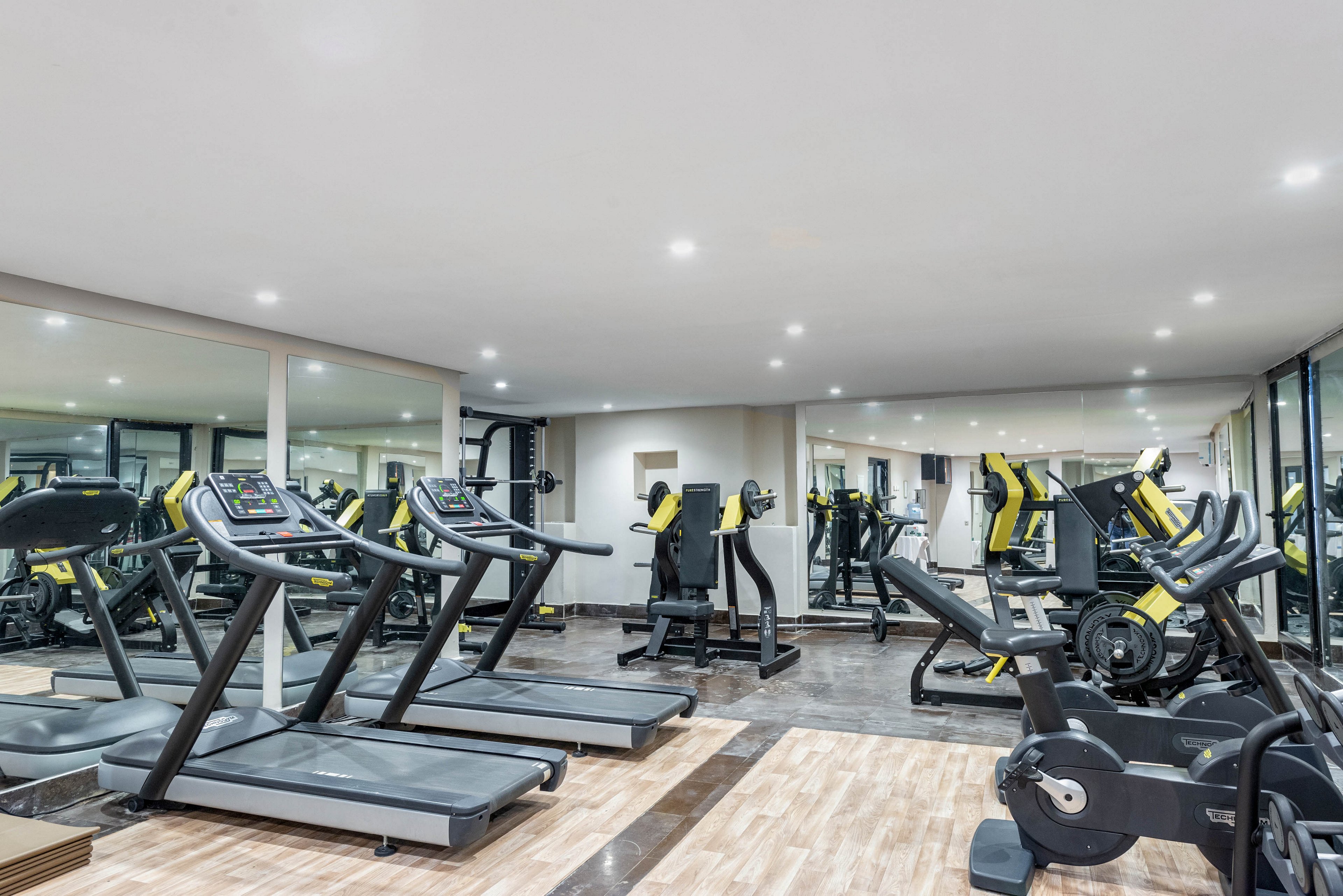 A spacious, modern gym interior features multiple treadmills and elliptical machines neatly arranged in rows. The room is brightly lit with recessed ceiling lights and has large mirrors along the walls, creating a sense of openness. Yellow and black accents on the fitness machines add a contemporary, high-energy feel. The polished floor and clean, uncluttered layout suggest a professional, well-maintained workout space.