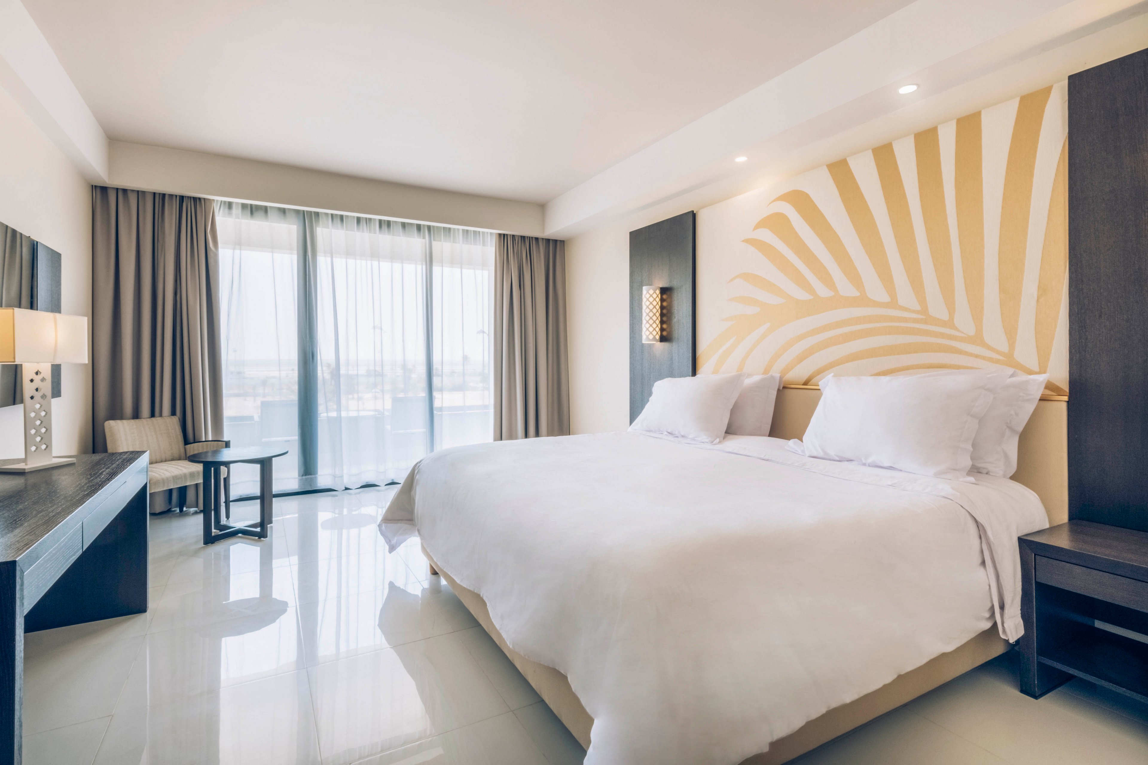 A spacious modern hotel bedroom features a large neatly made bed with crisp white linens. Floor-to-ceiling windows with sheer curtains open to a balcony and an outdoor view. Minimalist furniture, including a desk, side tables, and a small seating area, complements the neutral color palette. Soft lighting and a large abstract wall mural create a calm, contemporary atmosphere ideal for relaxation.