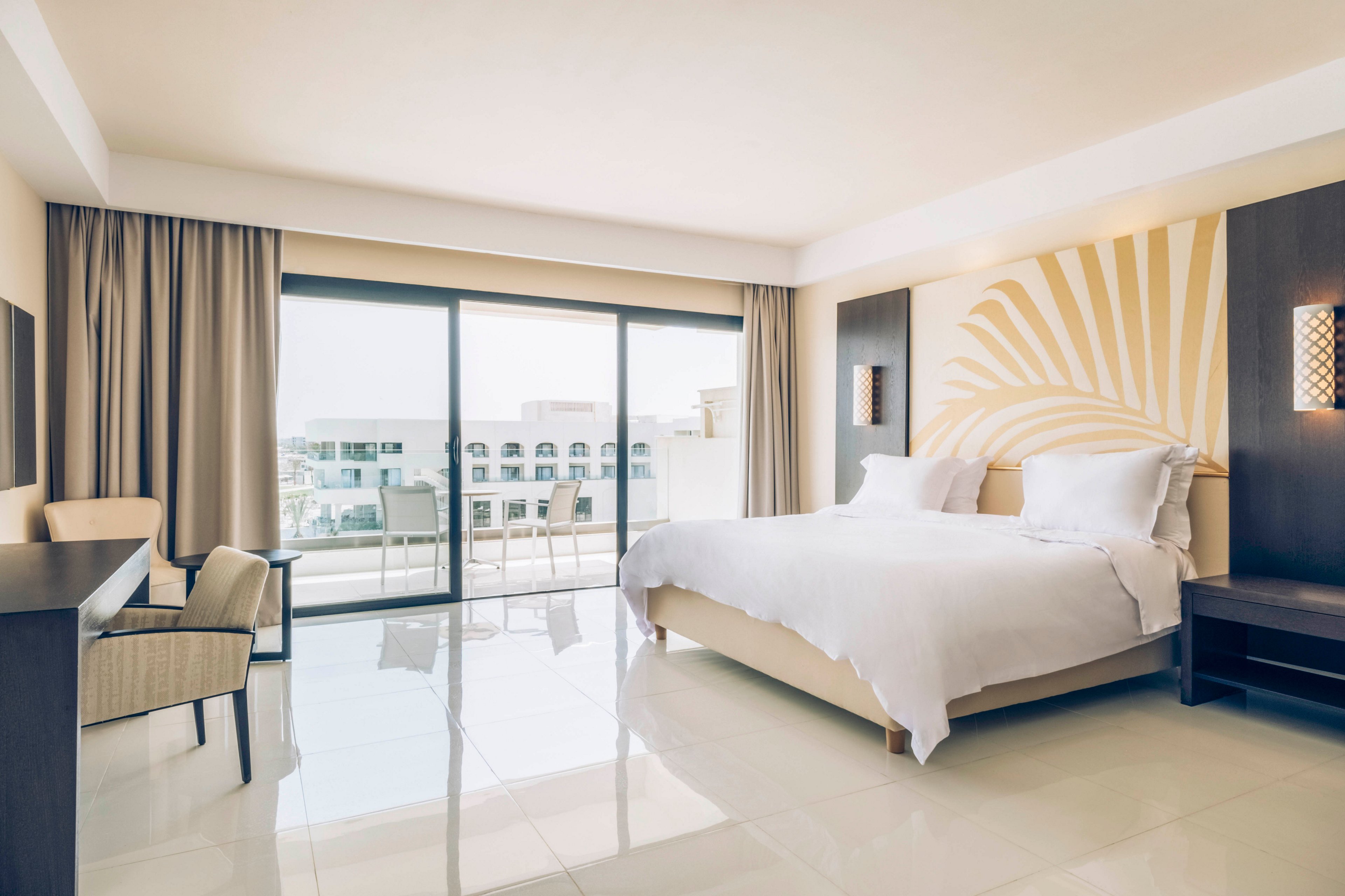 A spacious, modern hotel room features a large neatly made bed with white linens and a decorative headboard. Floor-to-ceiling sliding glass doors open to a balcony overlooking other resort buildings. Neutral tones, soft lighting, and minimalist furniture create a calm, contemporary atmosphere. The glossy tiled floor and natural daylight enhance the airy, clean feel of the space.
