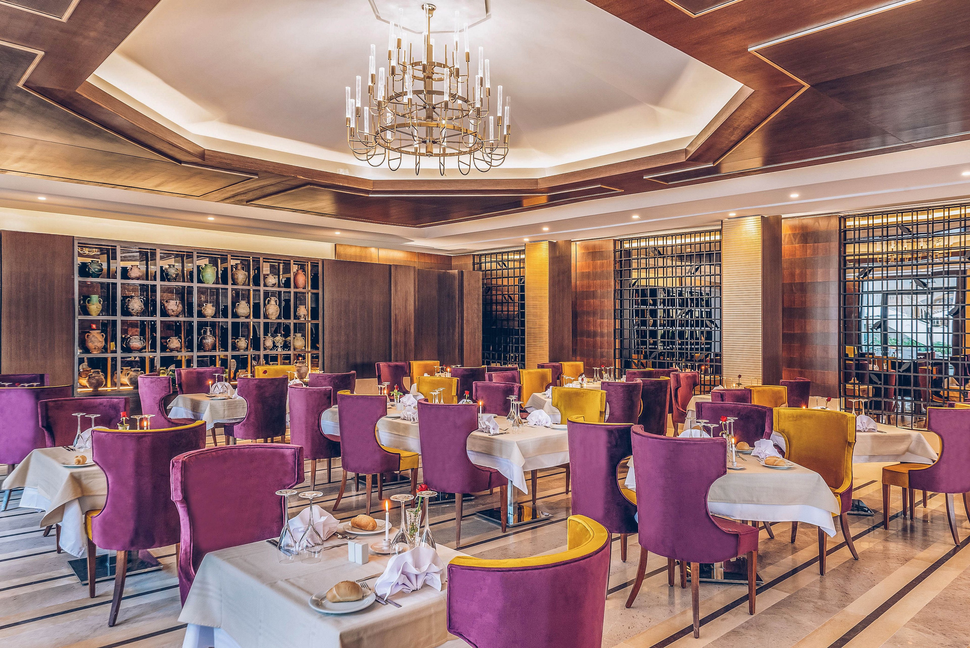 Upscale restaurant interior featuring neatly set dining tables with folded napkins and glassware. The room is furnished with vibrant purple and mustard yellow upholstered chairs around white tablecloths. Warm wooden wall panels and decorative shelving with vases and bottles create a sophisticated backdrop. A large chandelier and recessed ceiling lighting add a soft, inviting glow to the spacious, empty dining area.
