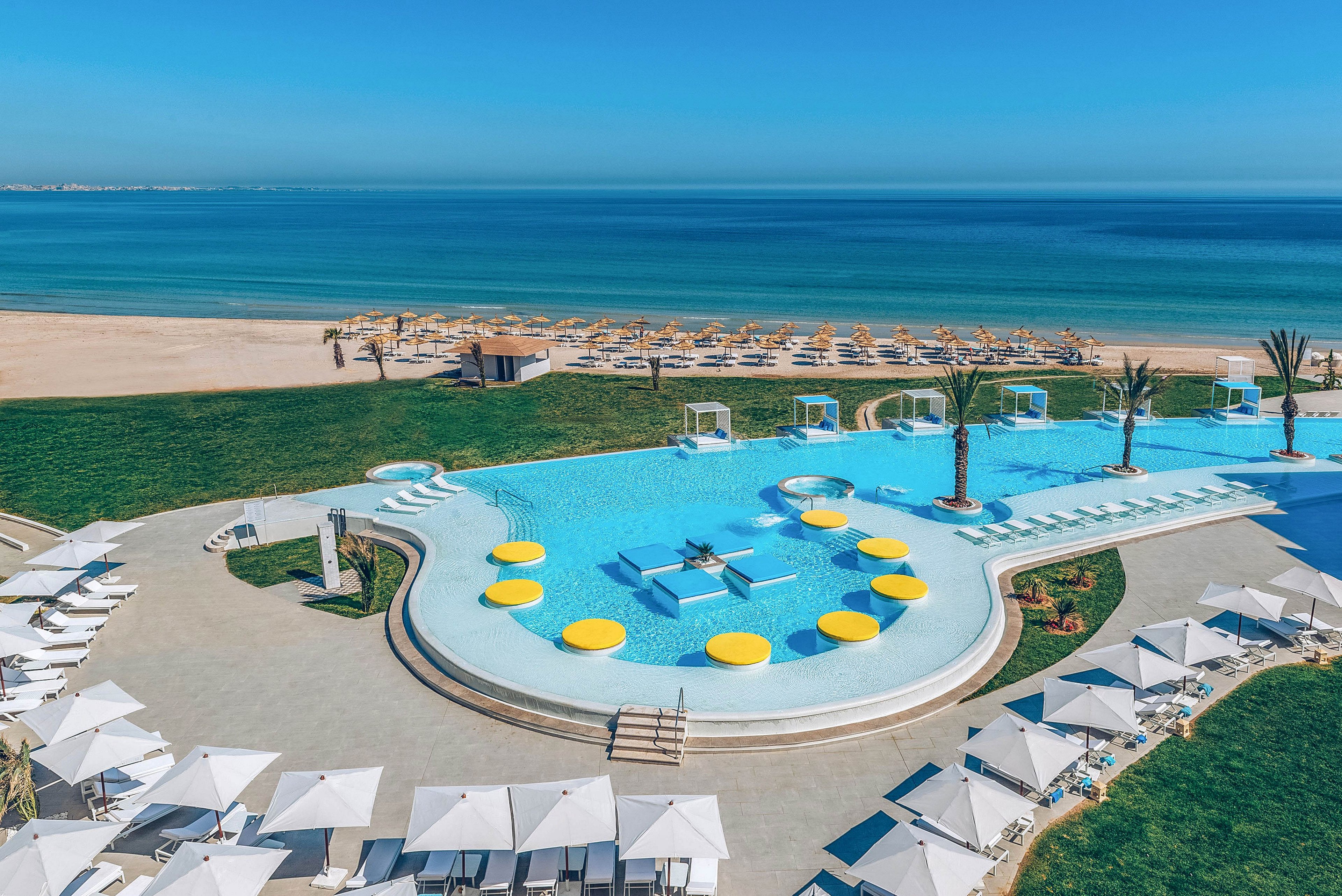 A large resort-style infinity pool overlooks a calm, turquoise sea and sandy beach. White sun loungers and umbrellas surround the pool, creating a clean, modern leisure setting. Bright yellow circular floats and pool islands add playful contrast to the blue water. The clear sky and empty seating suggest a tranquil, relaxing vacation atmosphere.