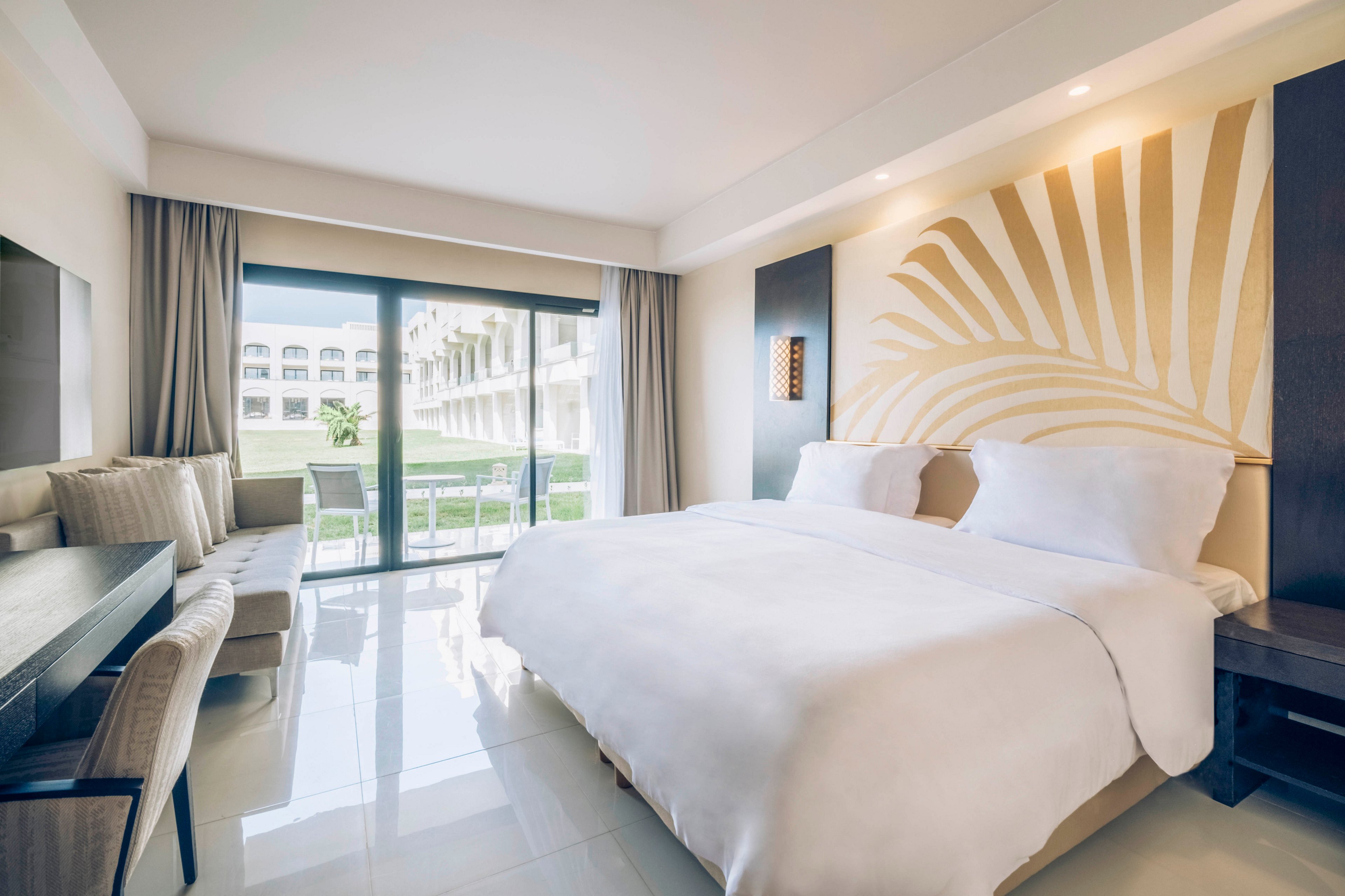 A spacious, modern hotel room features a large neatly made bed with white linens as the focal point. The room includes a sofa seating area, a desk, and floor-to-ceiling glass doors leading to a balcony. Soft neutral tones and a large abstract wall art create a calm, contemporary atmosphere. Outside, the balcony overlooks a green courtyard and adjacent resort buildings under natural daylight.