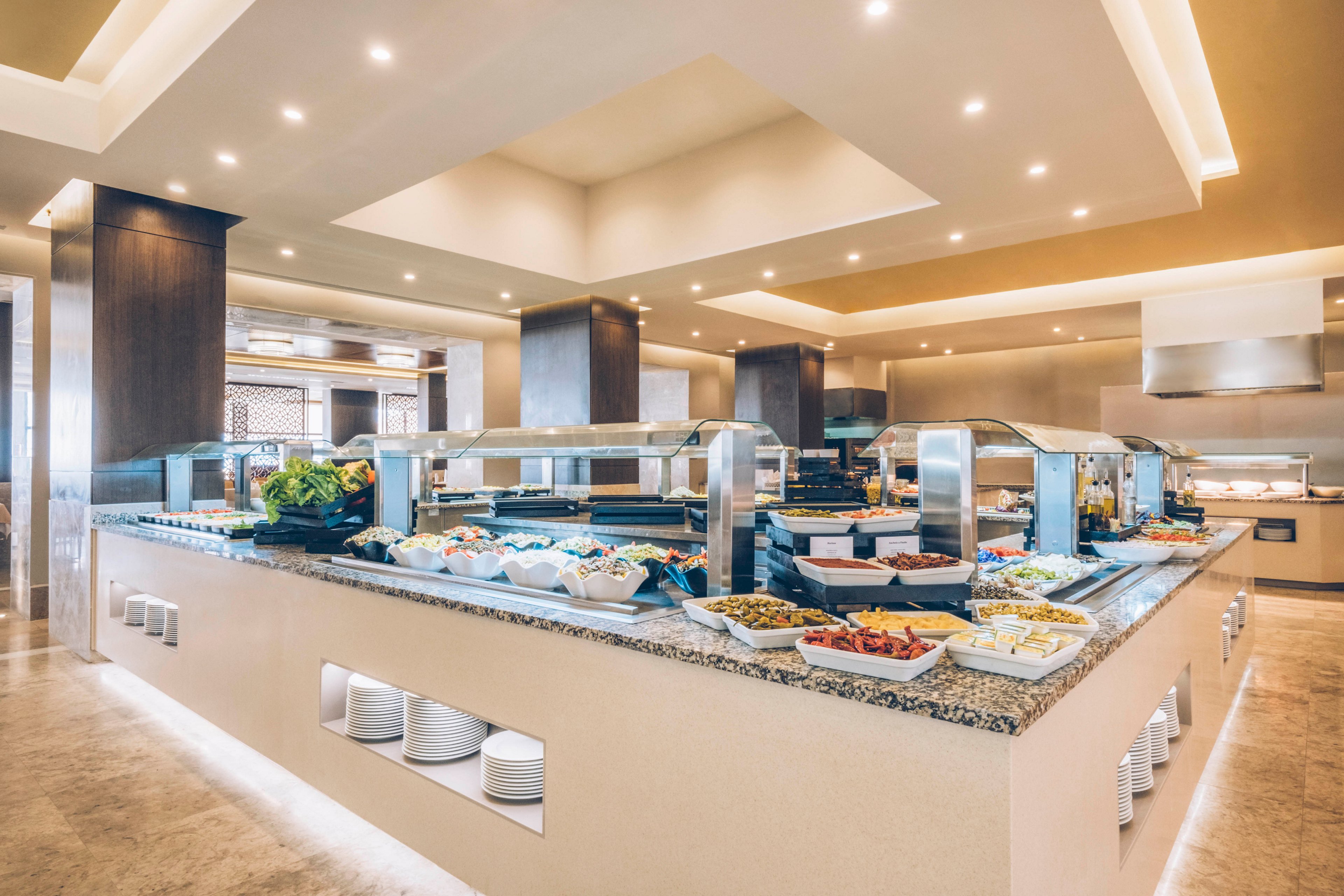 A spacious self-service buffet counter is filled with a variety of fresh salads, fruits, and hot dishes. The setting appears to be a modern hotel or resort restaurant with bright recessed ceiling lights. Granite countertops and glass sneeze guards frame the food displays, while stacks of white plates are neatly stored below. The overall atmosphere is clean, organized, and inviting for guests.