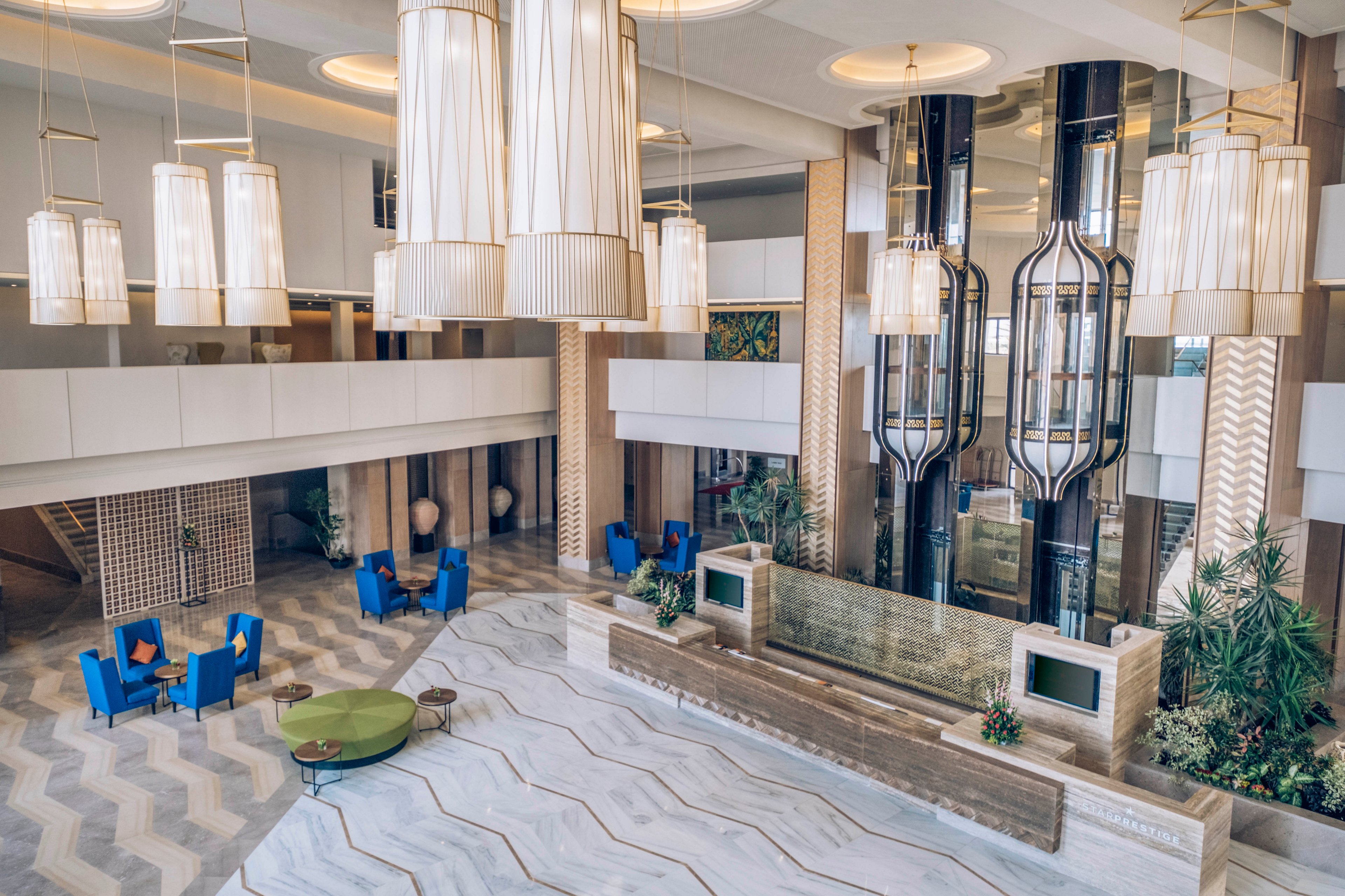 Spacious contemporary hotel lobby featuring large hanging chandeliers and a sleek reception desk. The setting includes patterned flooring, tall columns, and glass elevators framed by decorative metalwork. Blue lounge chairs with accent pillows and a round green table create inviting seating areas. Neutral tones with gold and beige details give the space an upscale, modern atmosphere.