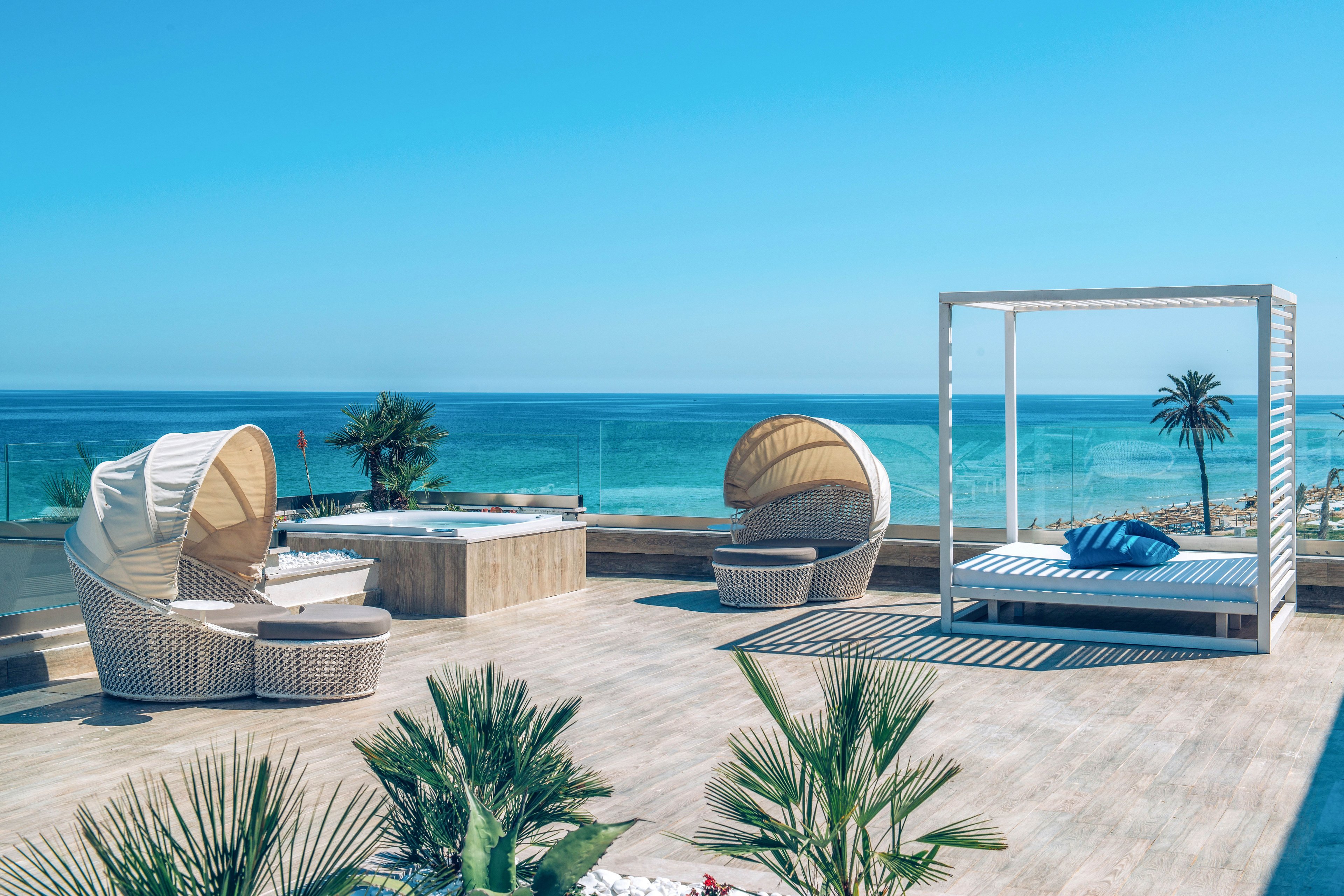 A modern rooftop terrace overlooks a bright blue ocean under a clear sky. Stylish wicker lounge chairs with curved canopies and a white four-poster daybed create an upscale relaxation area. A hot tub sits near the glass railing, enhancing the resort-like atmosphere. Green palm plants add a tropical touch to the clean, minimalist wooden deck.