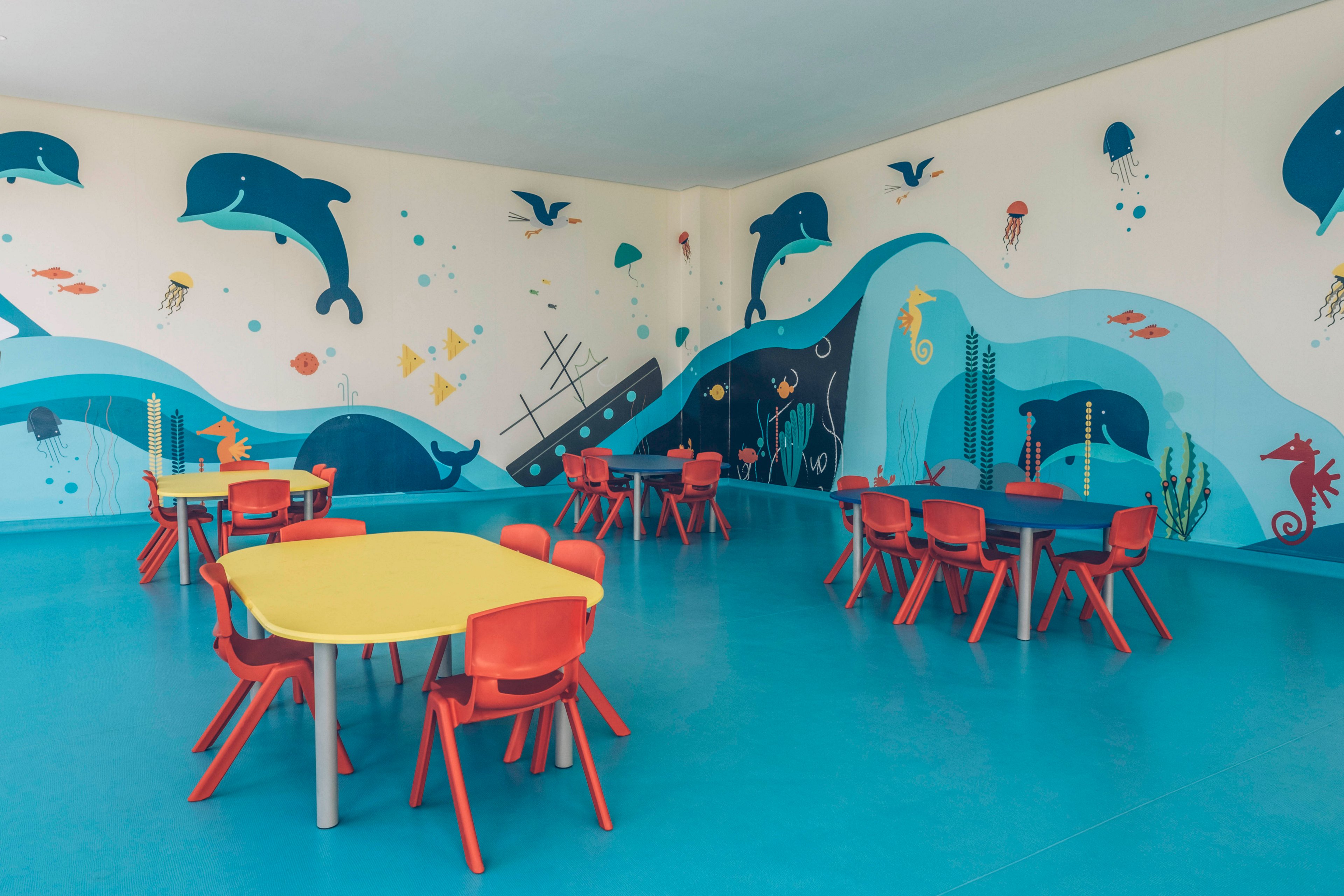 Brightly colored child-sized tables and chairs are arranged in an empty classroom. The walls feature a large undersea mural with dolphins, fish, jellyfish, and sea plants. The floor is a solid blue that complements the ocean theme on the walls. The space appears clean, modern, and inviting for young children.
