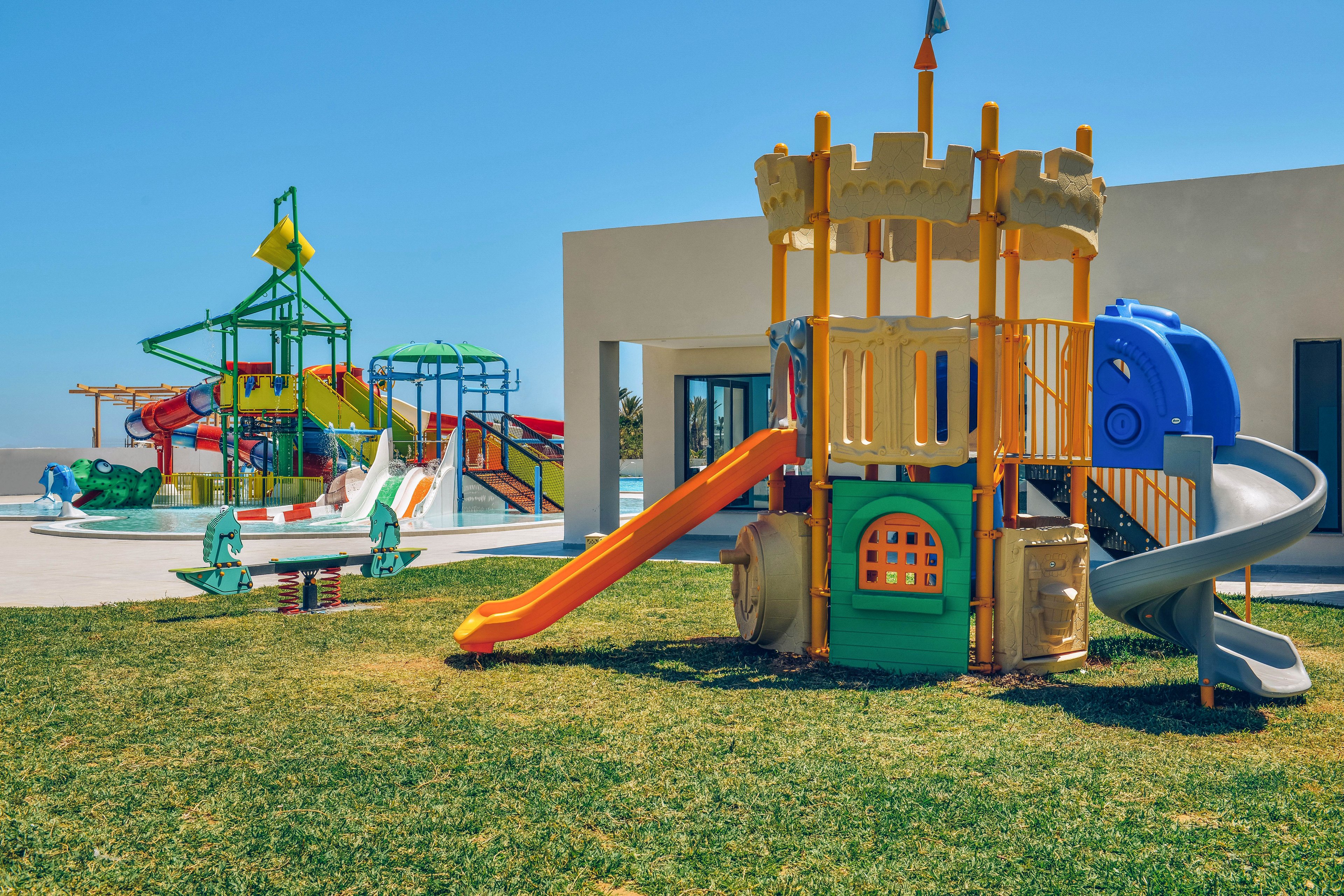 A brightly colored playground structure shaped like a castle stands on a grassy area under a clear blue sky. Multiple slides, climbing elements, and play panels are visible, including an orange slide and blue tunnel. In the background, additional playground equipment with water features and shade structures can be seen near a modern building. The scene suggests a sunny day at a family-friendly recreational area or resort.