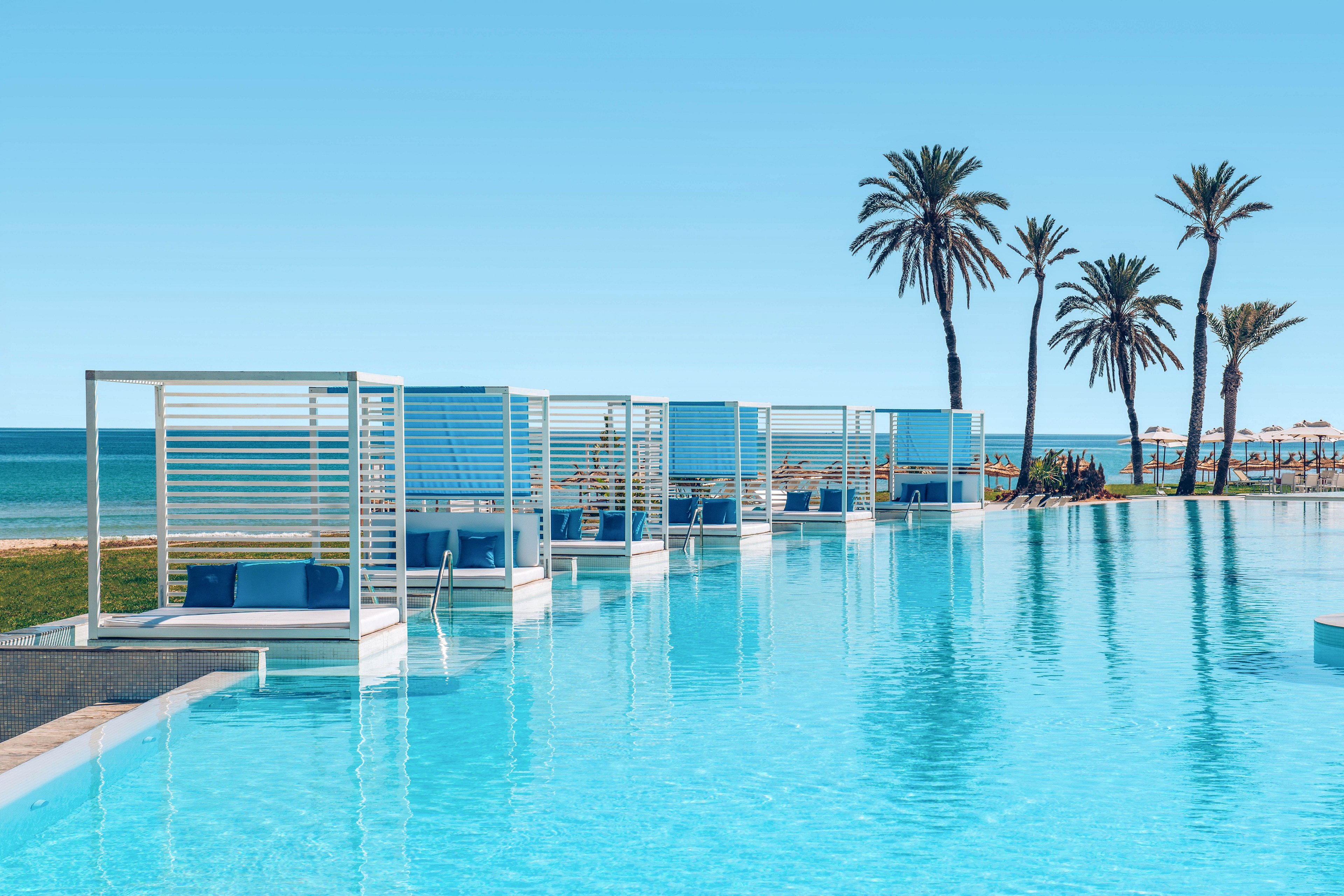 A luxurious infinity pool stretches toward the horizon beside the sea. Modern white and blue cabanas line the pool edge, offering shaded lounge areas. Tall palm trees rise in the background under a clear blue sky. The scene evokes a calm, upscale resort atmosphere perfect for relaxation and sunbathing.
