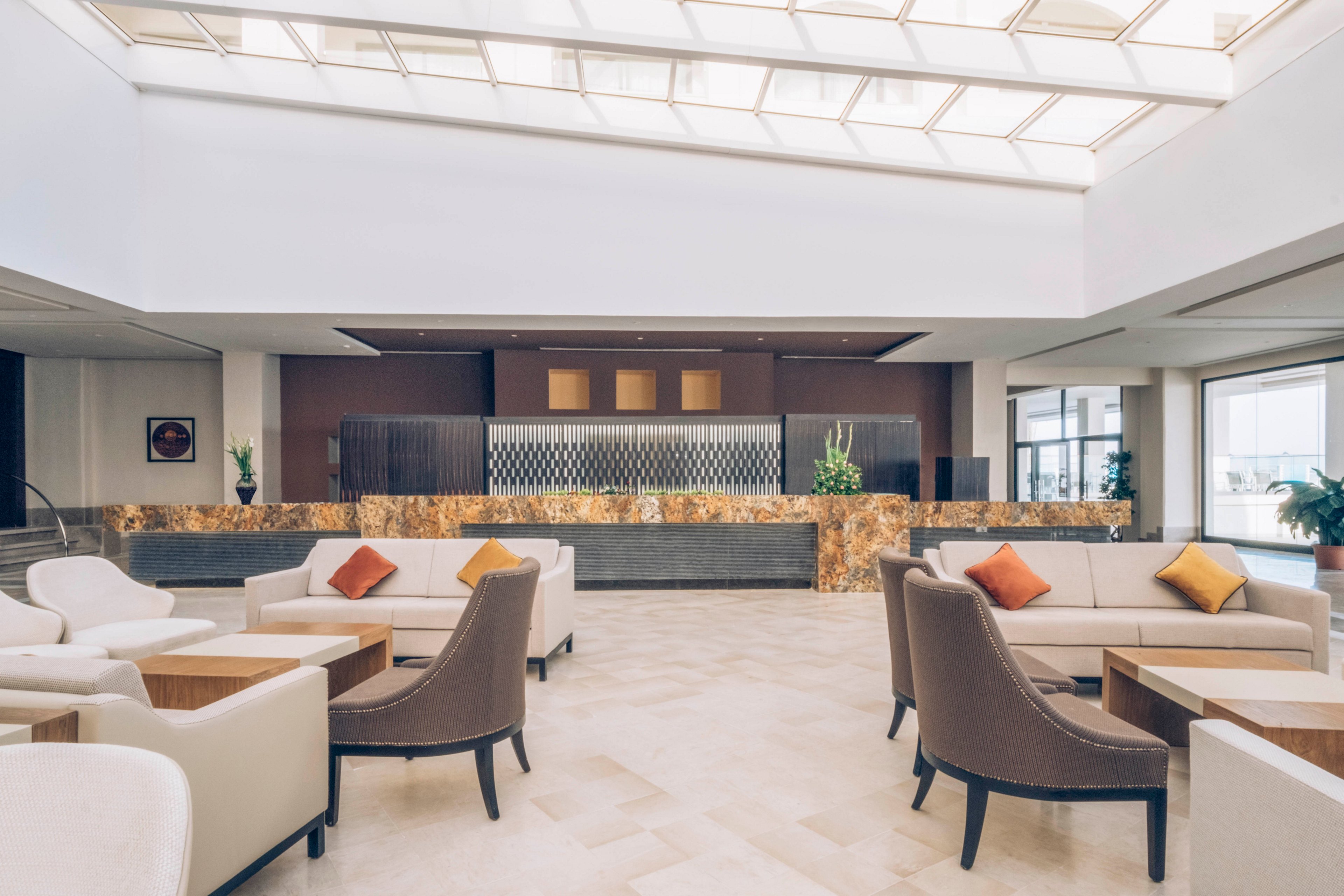 Spacious modern lobby interior featuring a central reception desk and contemporary lounge seating. Neutral-toned armchairs and sofas are accented with colorful throw pillows in orange, yellow, and red. Large windows and a skylight ceiling flood the space with natural light, highlighting the polished stone floor and marble-like counter. Decorative plants and clean architectural lines create a calm, upscale atmosphere suitable for hospitality or corporate environments.