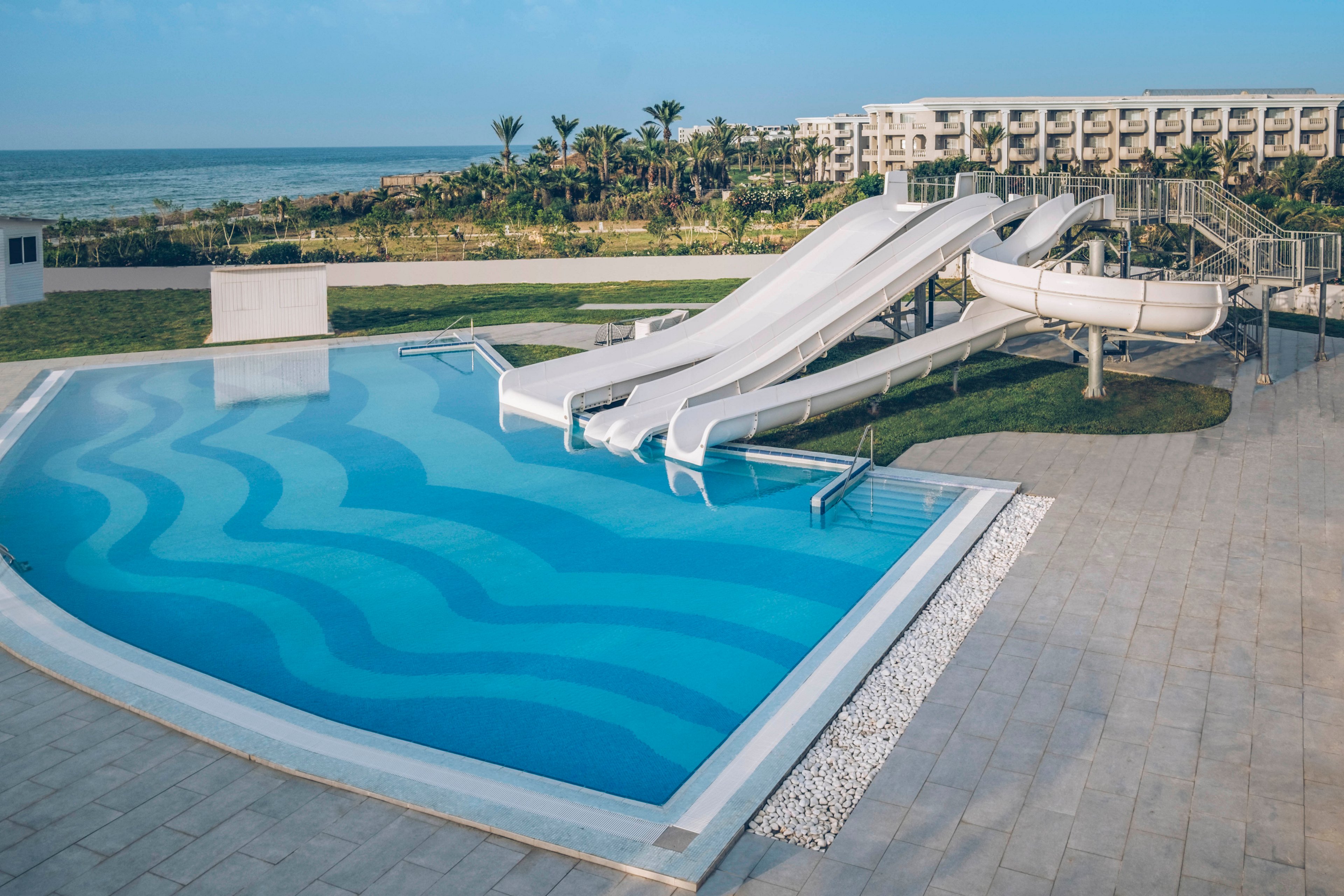 An outdoor resort swimming pool features multiple white water slides leading into clear blue water. The pool has decorative wave patterns on the floor and is surrounded by a tiled deck and small gravel border. In the background, palm trees, green lawns, and a large hotel building overlook the coastline and sea. The scene suggests a sunny, relaxing vacation destination focused on leisure and family fun.