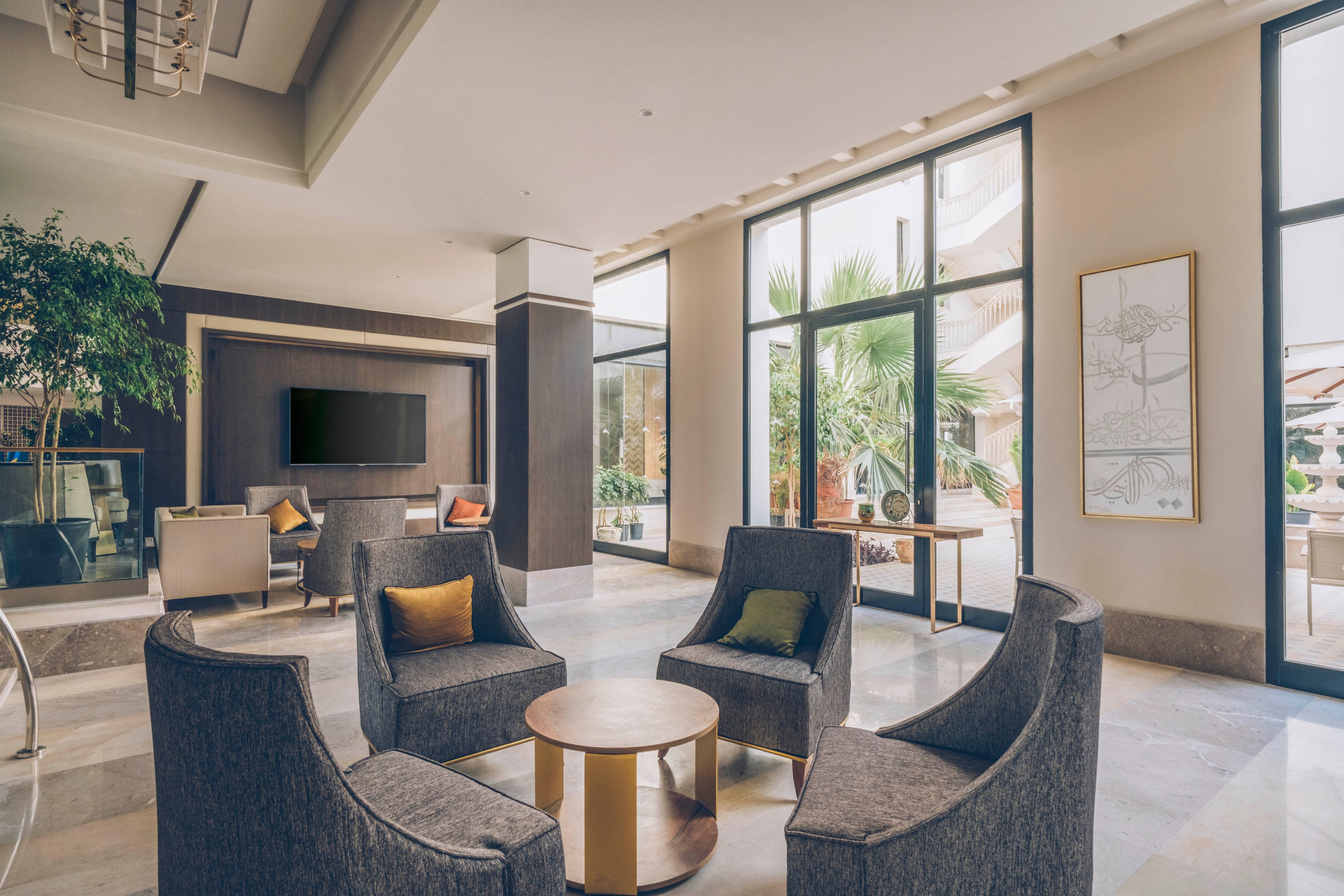 A contemporary hotel lobby lounge features a circular arrangement of gray armchairs around a small round coffee table. Large floor-to-ceiling windows let in natural light and reveal greenery and palm trees outside. Neutral tones dominate the interior, accented by colorful throw pillows and indoor plants. A wall-mounted television and framed artwork add to the upscale, relaxed atmosphere.