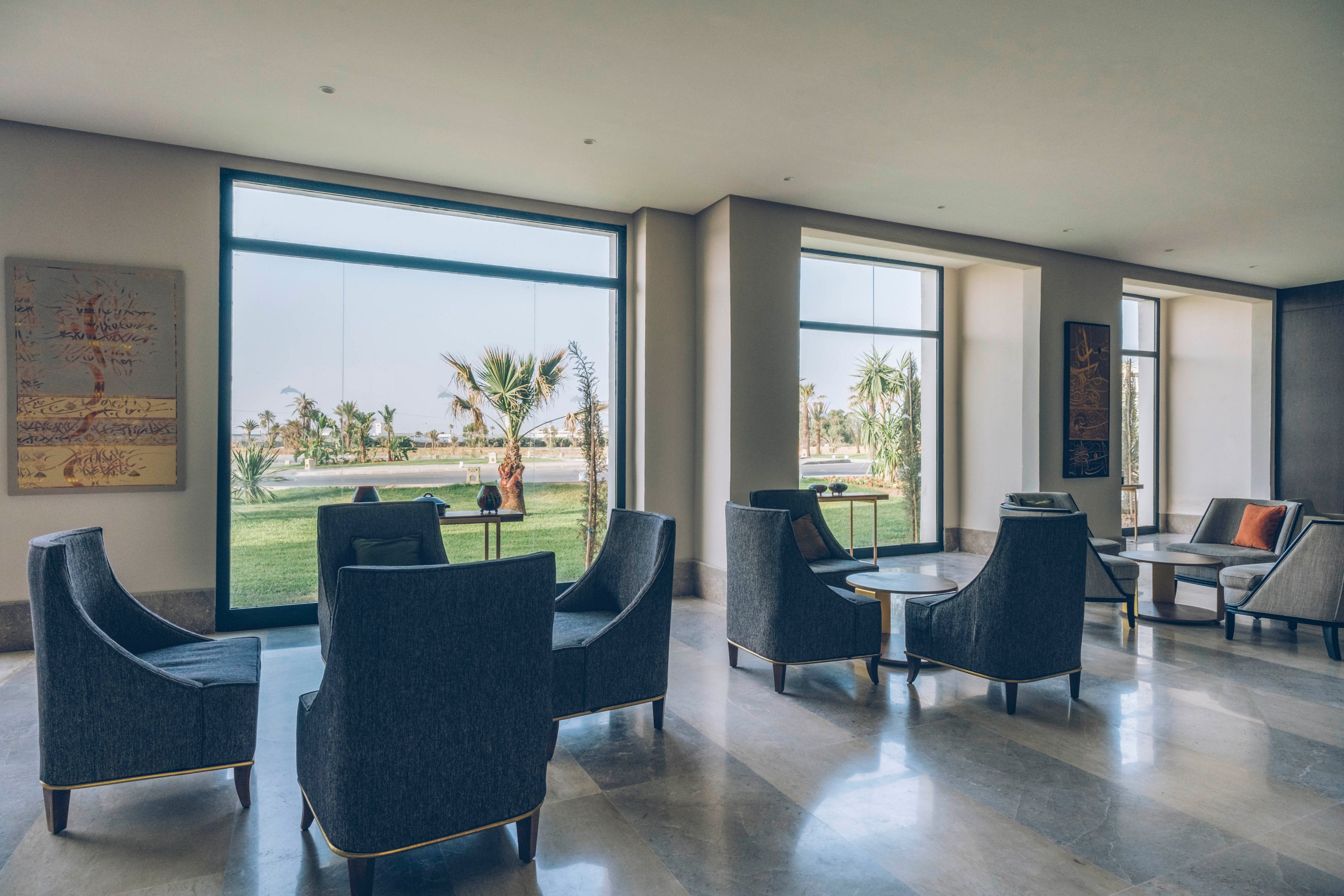 A contemporary lounge area features dark upholstered armchairs arranged around small tables. Large floor-to-ceiling windows open onto a green lawn with palm trees and outdoor seating. Soft natural light fills the minimalist interior, highlighting neutral walls and polished flooring. Framed artwork and accent cushions add subtle decorative touches to the calm, spacious setting.