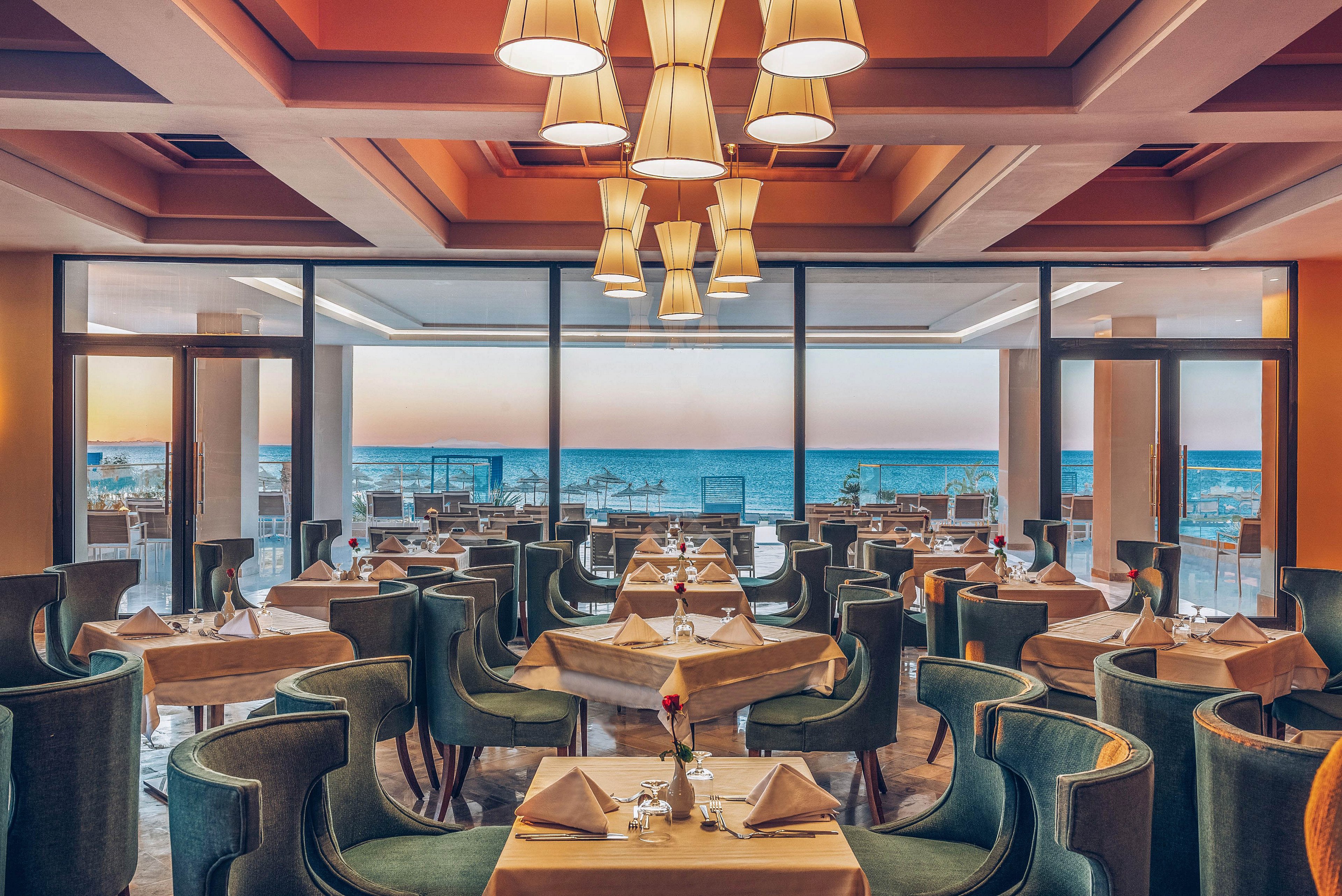 Upscale restaurant dining room with neatly arranged tables overlooks a calm ocean view at sunset. Plush green chairs surround square tables set with folded napkins, glassware, and single flower centerpieces. Large floor-to-ceiling windows frame the sea and pastel sky, creating an open, airy atmosphere. Warm pendant lights and a muted color palette add to the sophisticated, romantic ambiance.