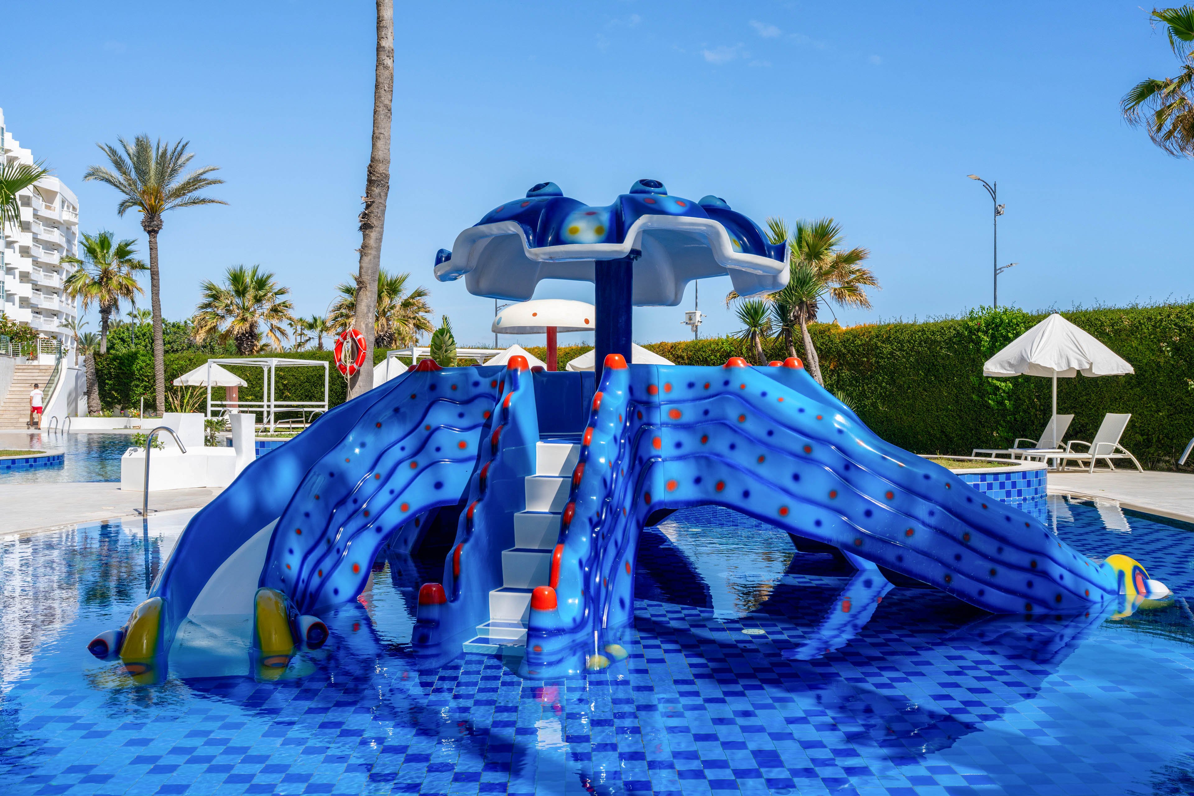 A bright blue octopus-shaped water slide stands in the middle of a resort swimming pool. The slide features multiple curved arms, a central staircase, and a mushroom-style canopy at the top. Palm trees, lounge chairs, and umbrellas surround the pool area under a clear blue sky. The scene suggests a family-friendly vacation destination with playful aquatic attractions.