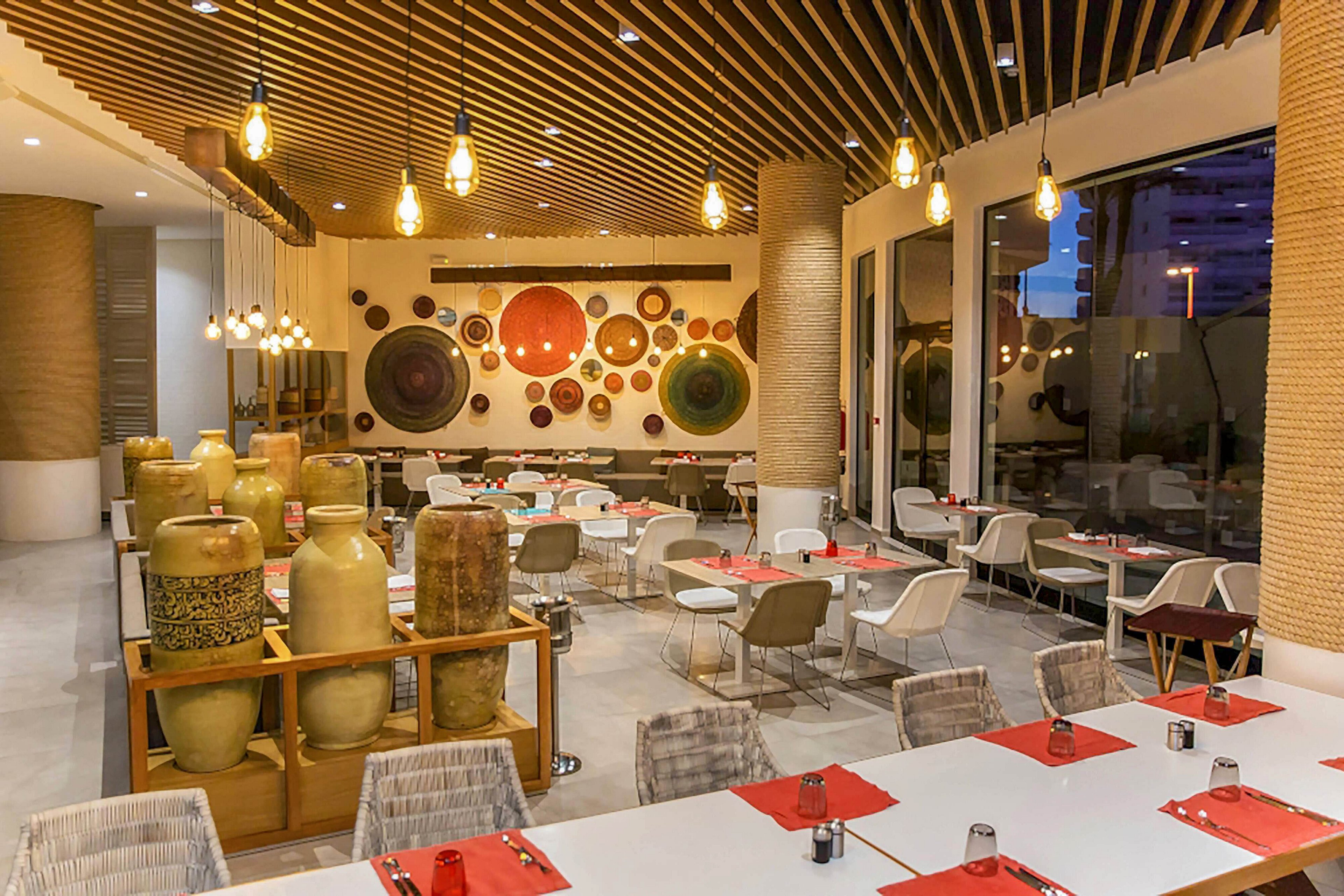 A contemporary restaurant dining area features neatly arranged tables with red placemats and white chairs. Large ceramic vases are displayed on stands near the entrance, adding an artistic touch. The back wall is decorated with an array of colorful circular plates in various sizes. Warm pendant lighting and a slatted wooden ceiling create a cozy, inviting atmosphere.