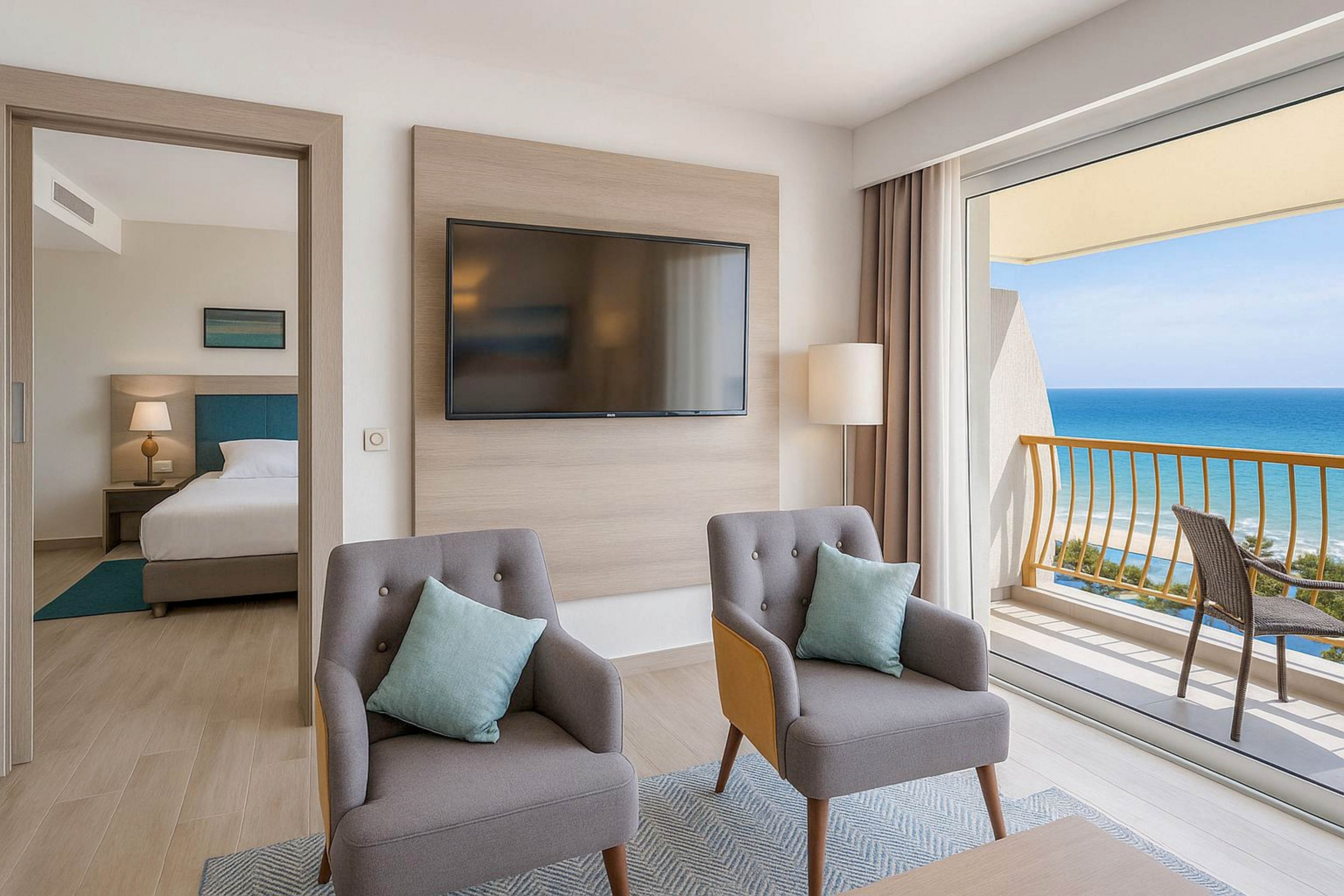 Contemporary hotel suite living area featuring two gray armchairs with pastel cushions and a wall-mounted flat-screen TV. A separate bedroom with a neatly made bed and bedside lamp is visible through an open doorway. Large floor-to-ceiling windows open onto a balcony with a railing overlooking a bright blue ocean and clear sky. Neutral tones and minimalist decor create a calm, relaxing vacation atmosphere.